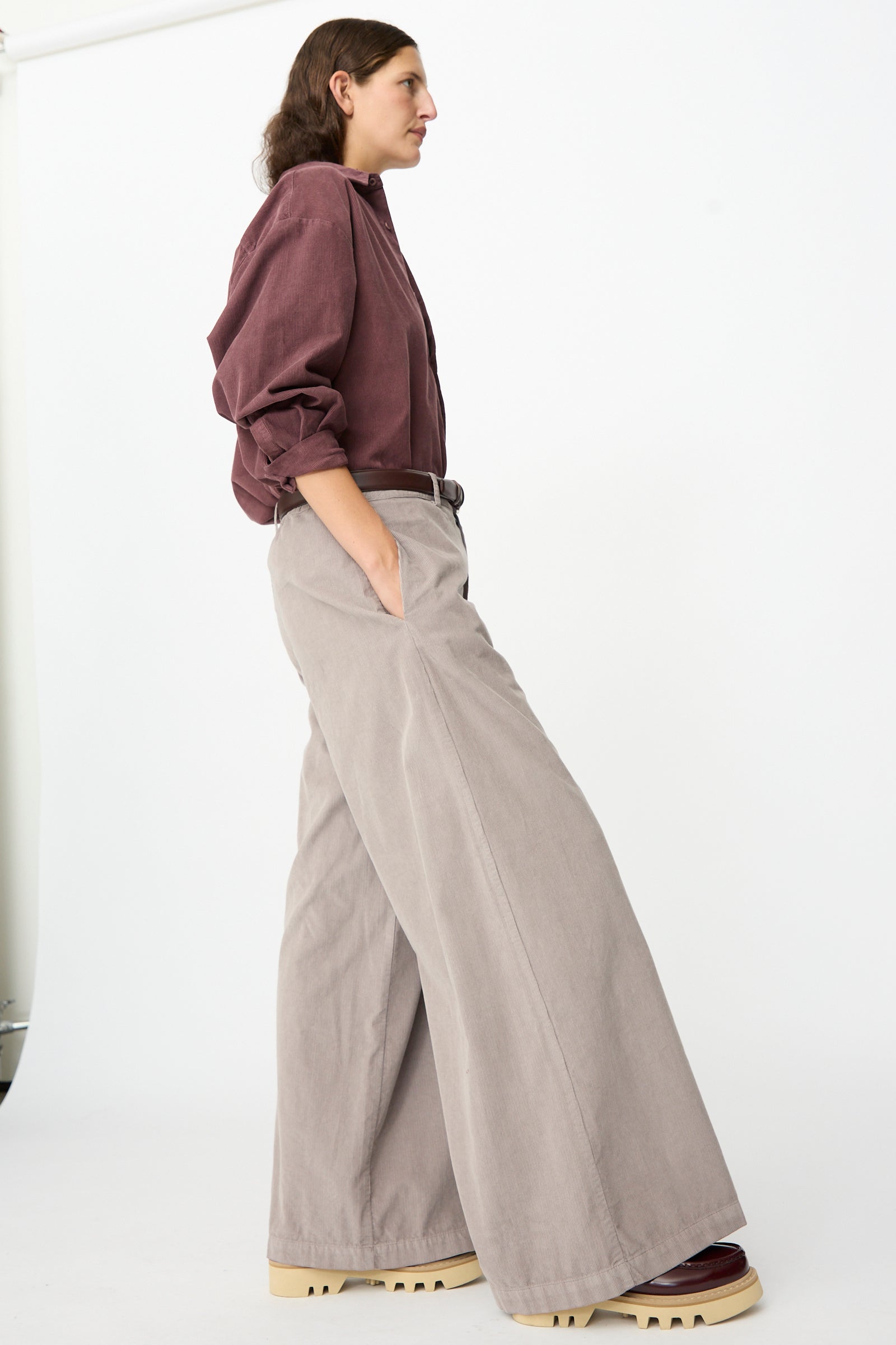 A person stands in profile against a white background, wearing the Labo.Art Binocchio Trouser in Malva—a wide-leg pant crafted from soft cotton needlecord—paired with a burgundy shirt, platform shoes, and hands casually tucked in pockets.