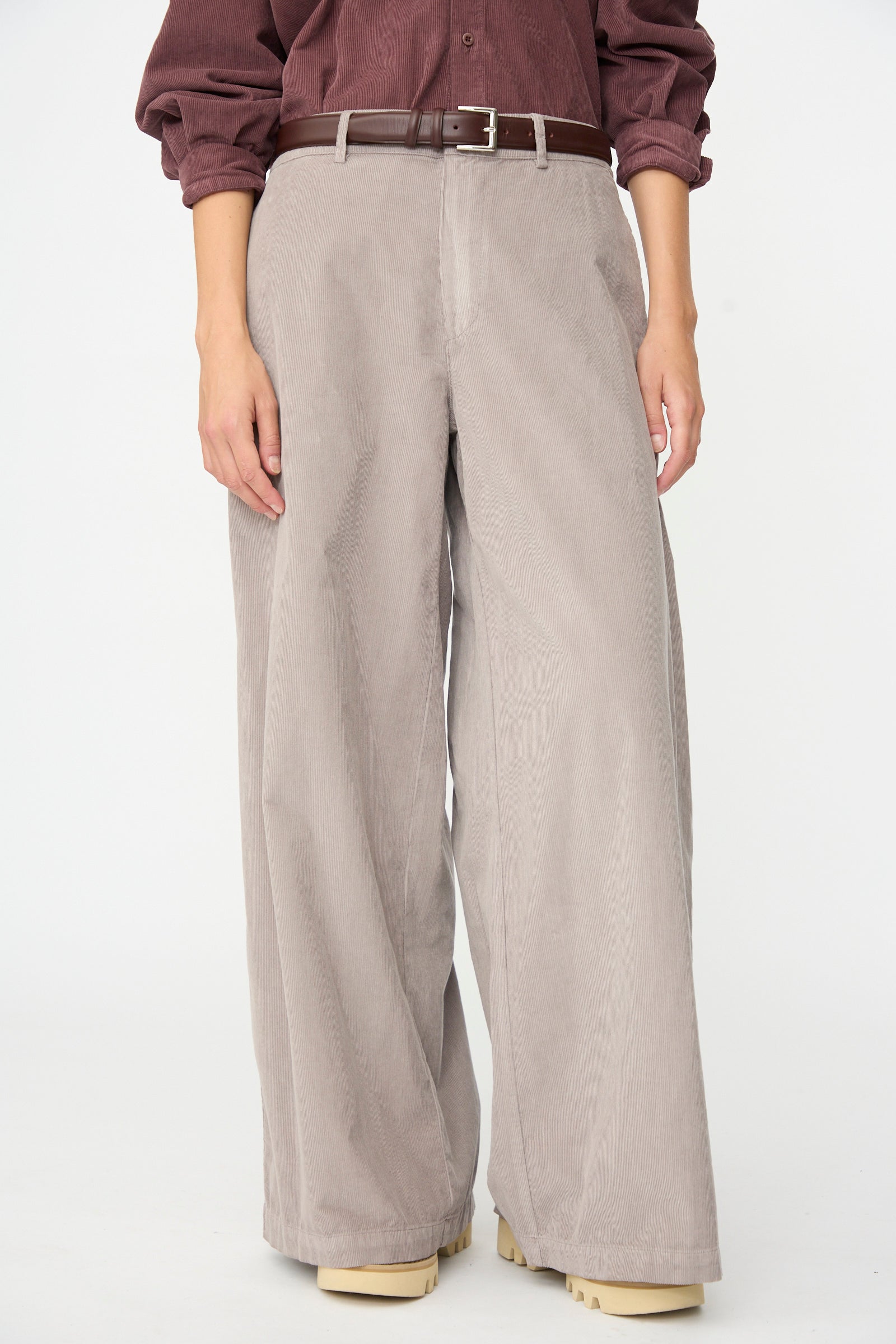 A person wearing the Labo.Art Binocchio Trouser in Malva—wide-leg beige cotton needlecord pants made in Italy—paired with a brown belt and a maroon shirt with rolled sleeves, stands against a plain white background.