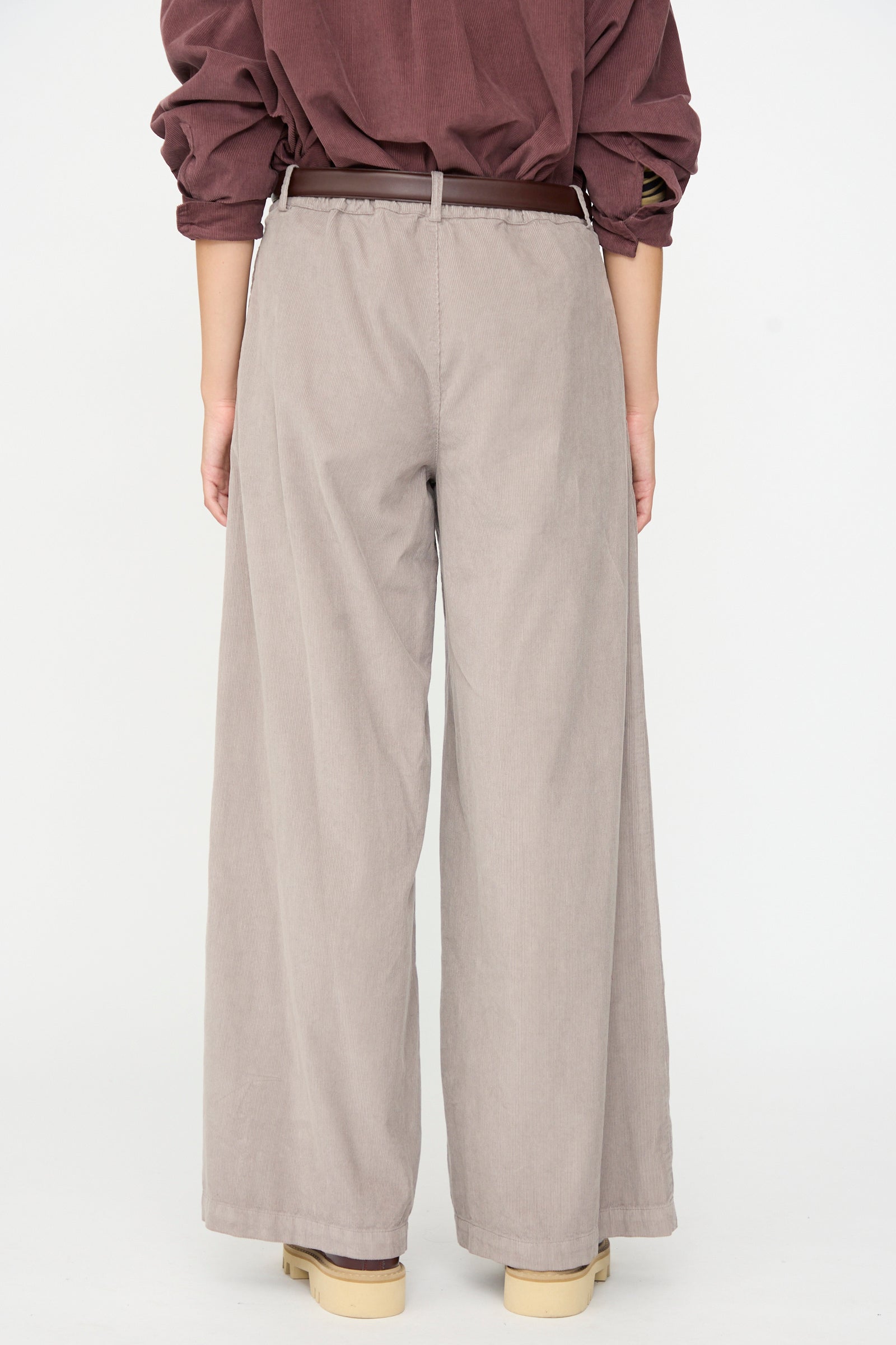 A person stands facing away against a plain white background, dressed in a brown long-sleeve shirt and Labo.Art’s Binocchio Trouser in Malva—beige wide-leg pants crafted from soft cotton needlecord—paired with a brown belt and beige shoes.