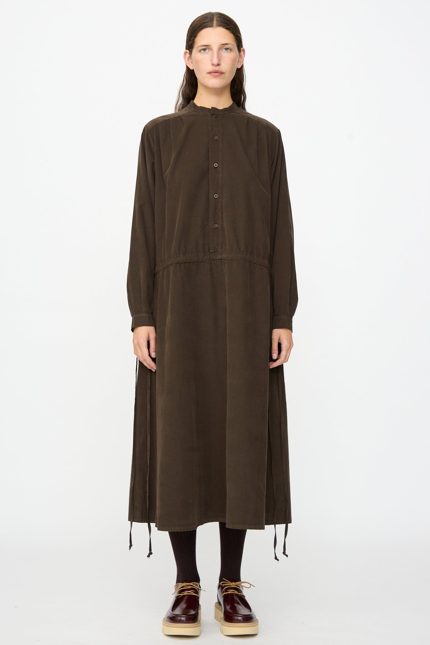 A person stands facing forward against a plain white background, wearing the Clio Dress in Cacao by Labo.Art—a long sleeve midi dress made from cotton needlecord fabric, styled with matching tights and shoes for a look that exudes comfort and timeless elegance.