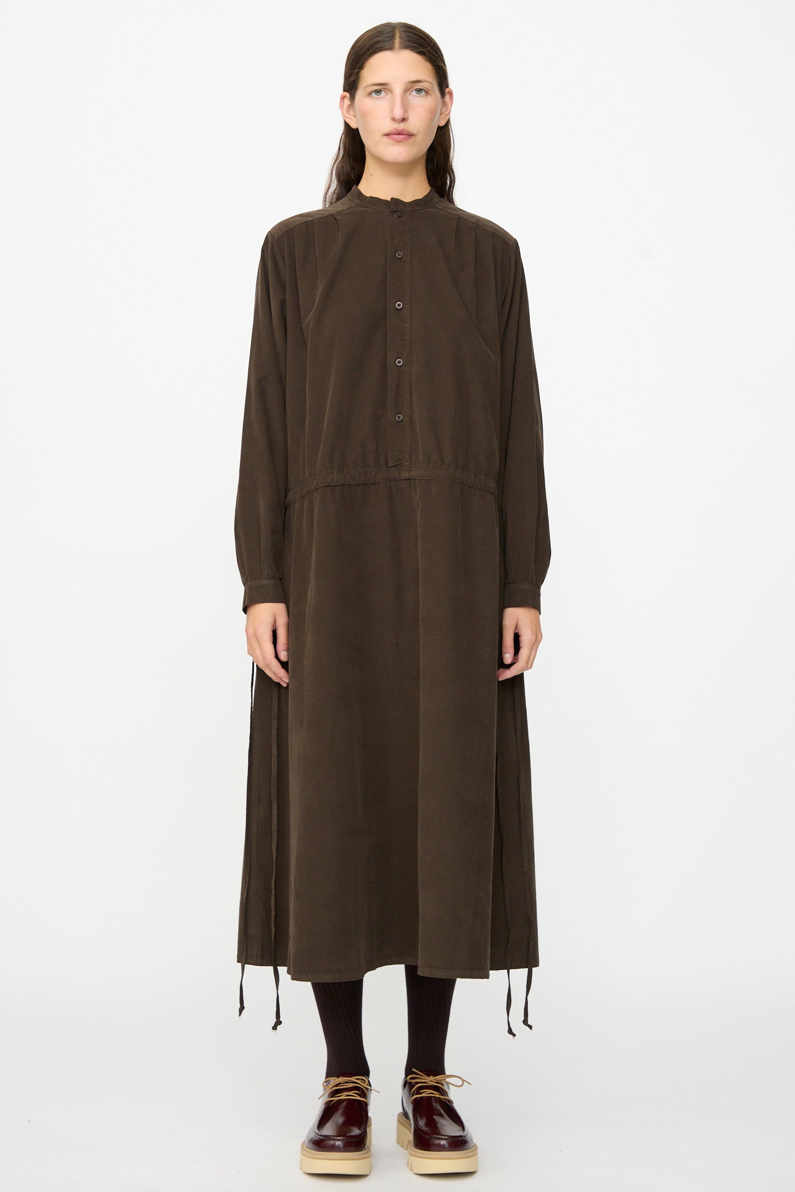 A person stands facing forward against a plain white background, wearing the Clio Dress in Cacao by Labo.Art—a long sleeve midi dress made from cotton needlecord fabric, styled with matching tights and shoes for a look that exudes comfort and timeless elegance.
