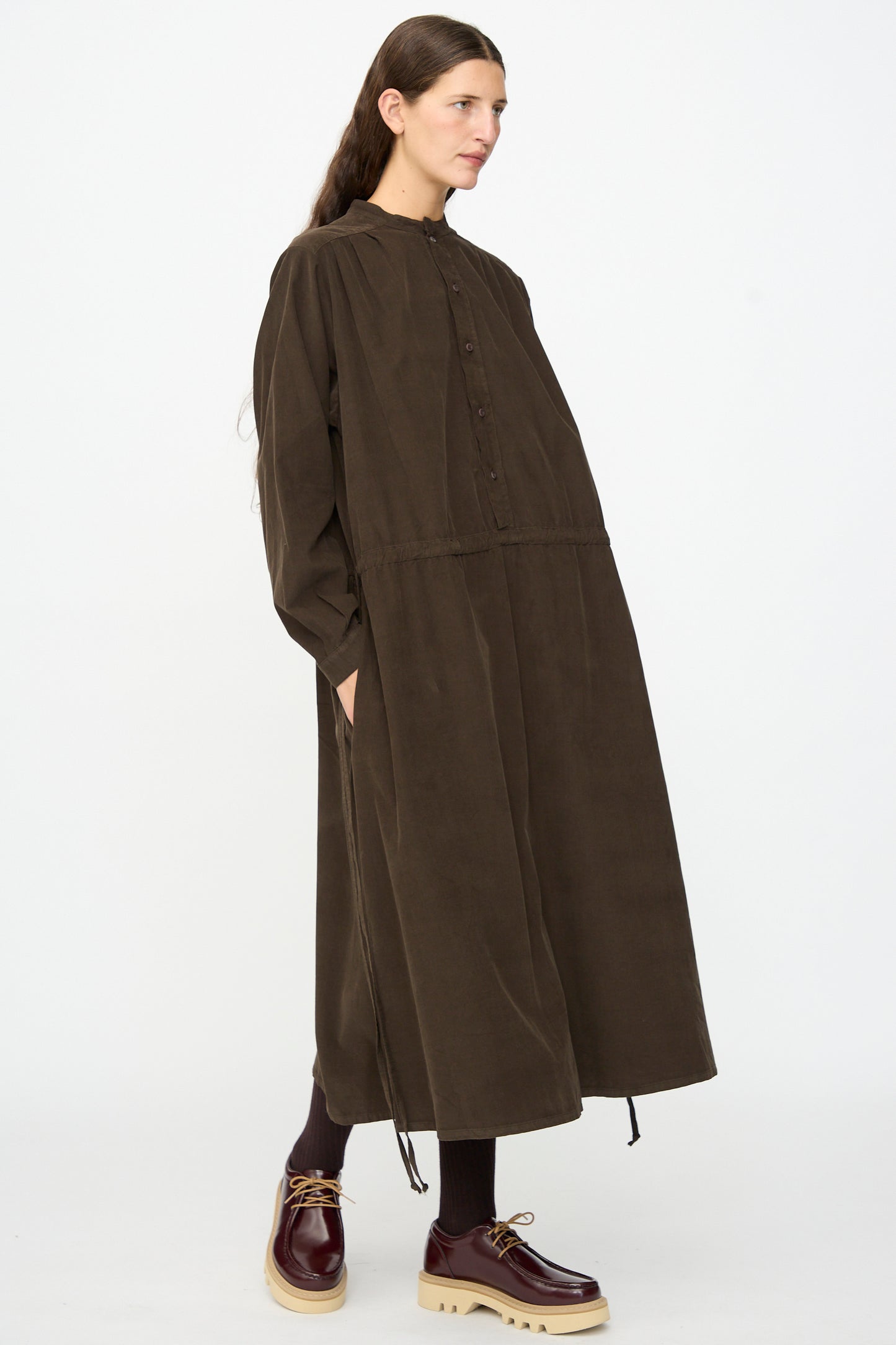 A woman stands against a plain white background wearing the Clio Dress in Cacao by Labo.Art—a long, loose-fitting brown cotton needlecord midi dress with buttons and pockets. She pairs it with brown socks and chunky brown shoes. Made in Italy, this long sleeve dress blends comfort and effortless style.