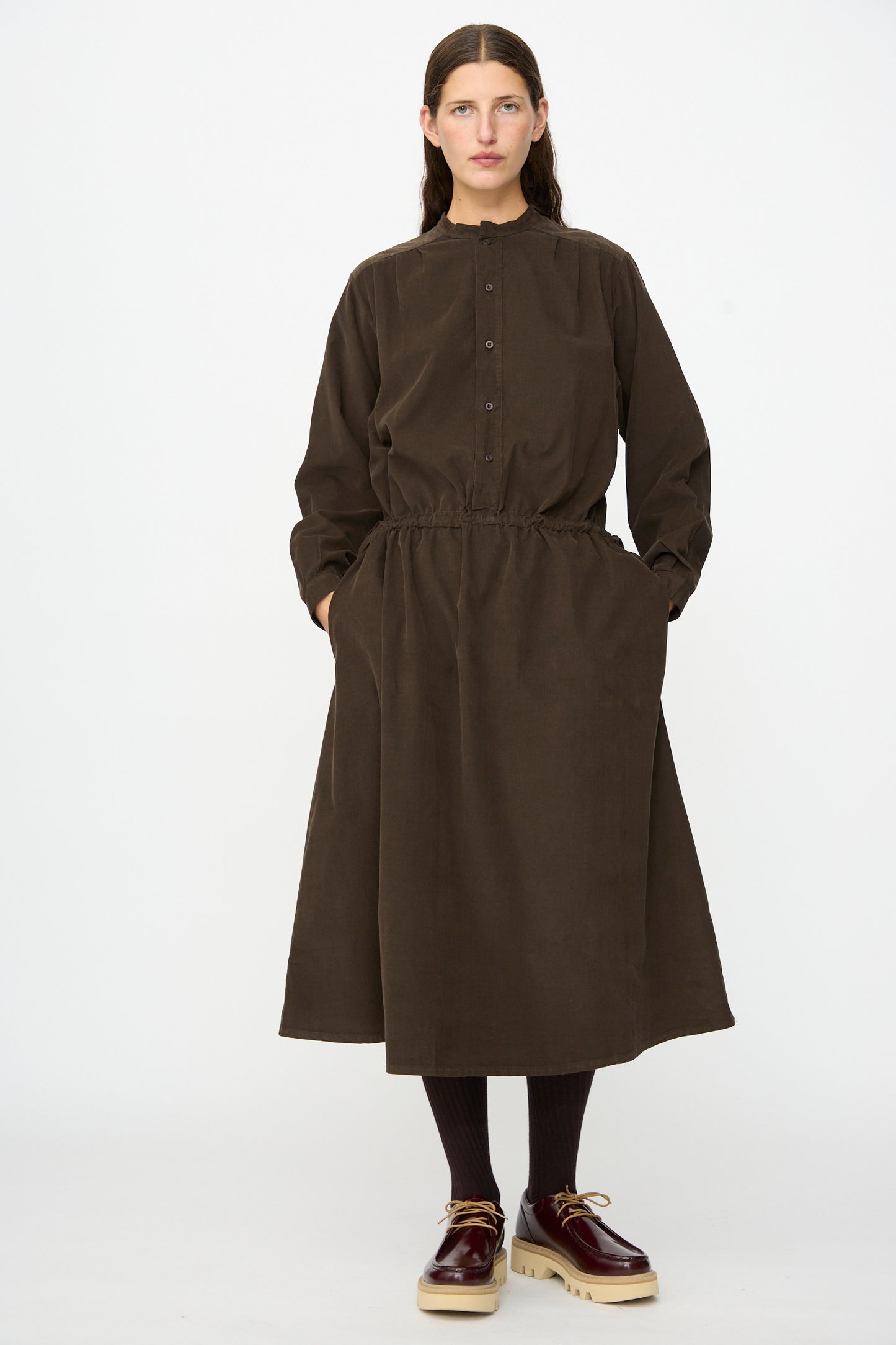 A person stands against a white background wearing the Labo.Art Clio Dress in Cacao—a long sleeve, button-up cotton needlecord midi dress with pockets—paired with brown socks and brown platform shoes.