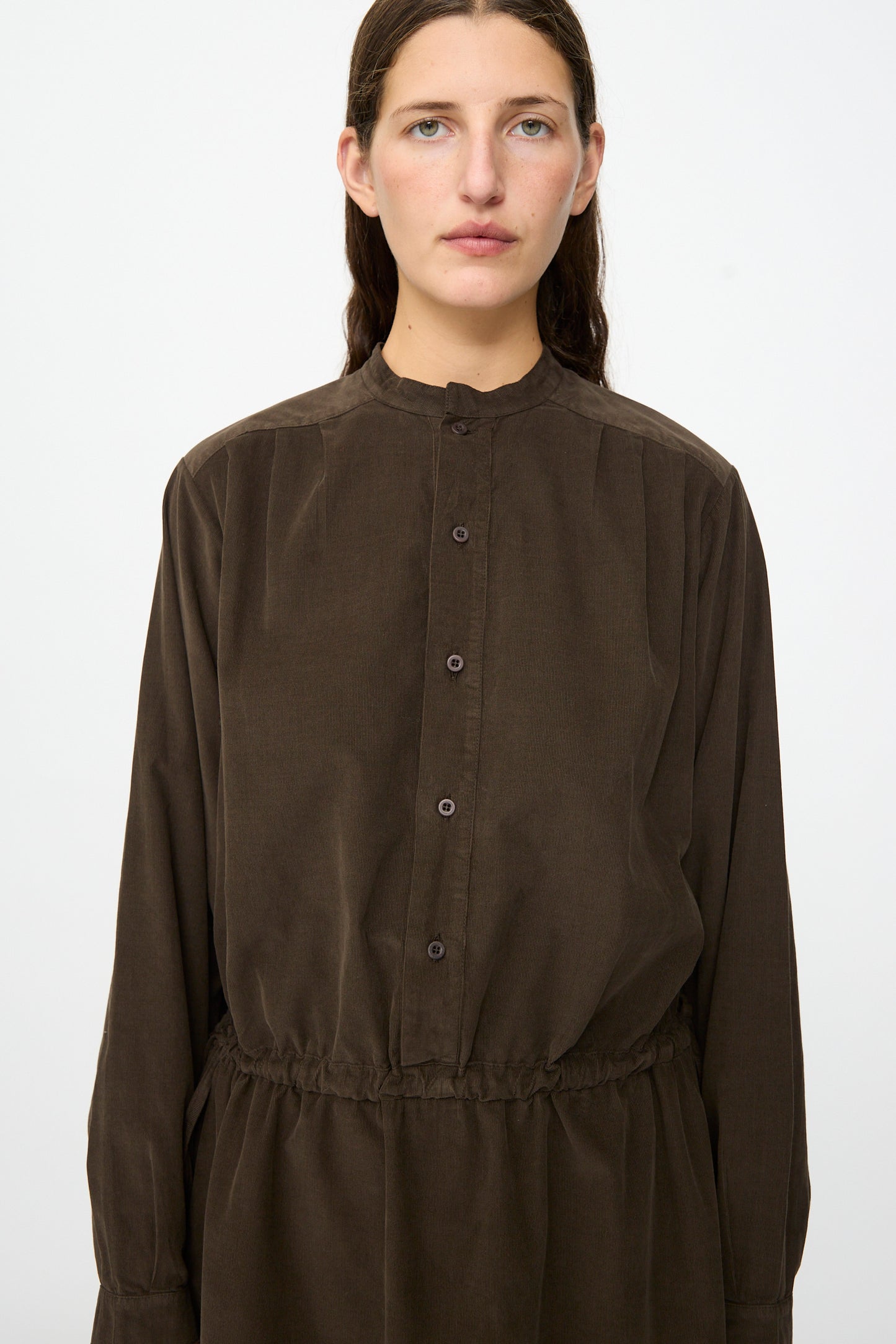 A person with long brown hair wears the Clio Dress in Cacao by Labo.Art—a dark brown, cotton needlecord midi dress with long sleeves and a gathered waist—standing against a plain white background. This dress is made in Italy.