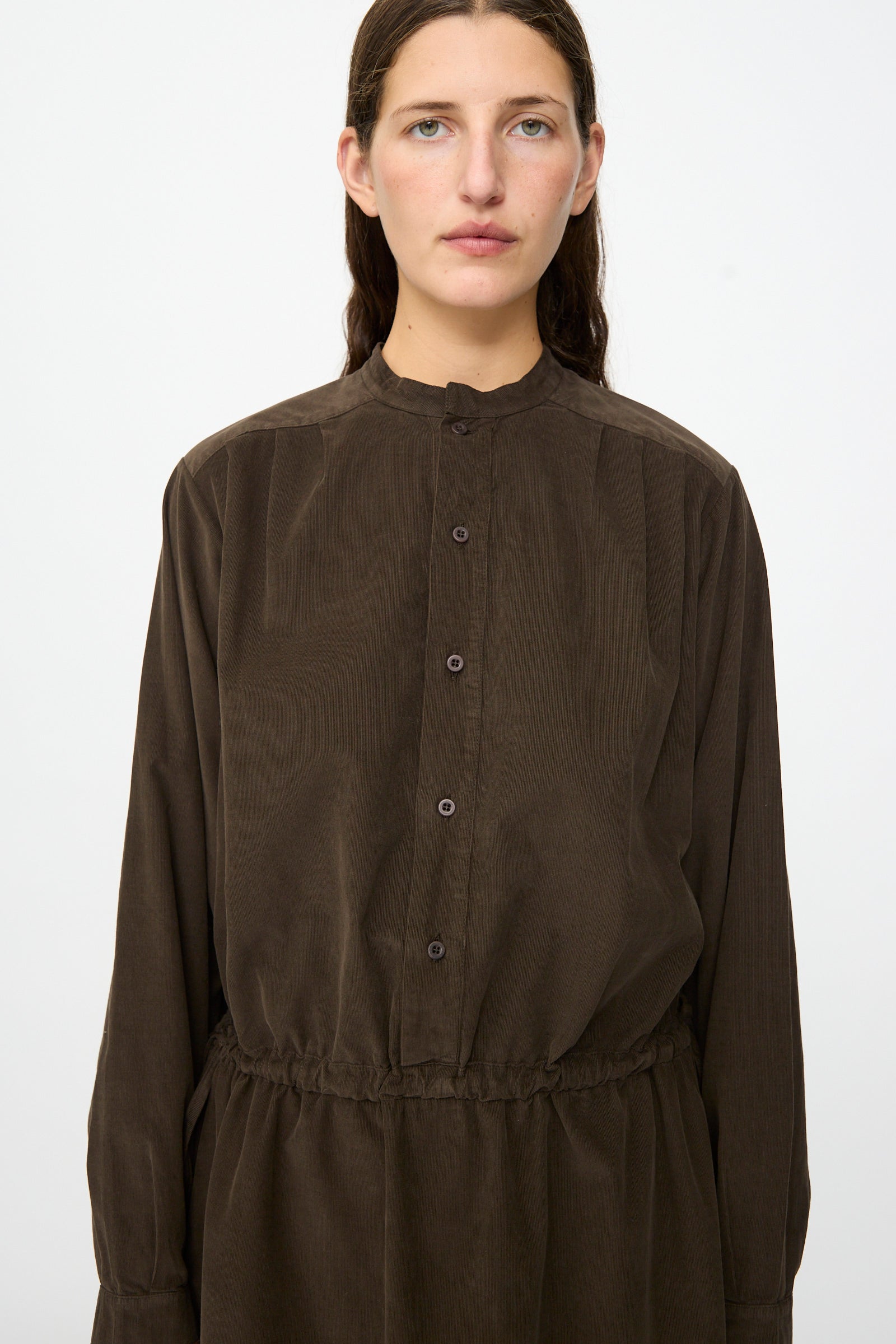 A person with long brown hair wears the Clio Dress in Cacao by Labo.Art—a dark brown, cotton needlecord midi dress with long sleeves and a gathered waist—standing against a plain white background. This dress is made in Italy.