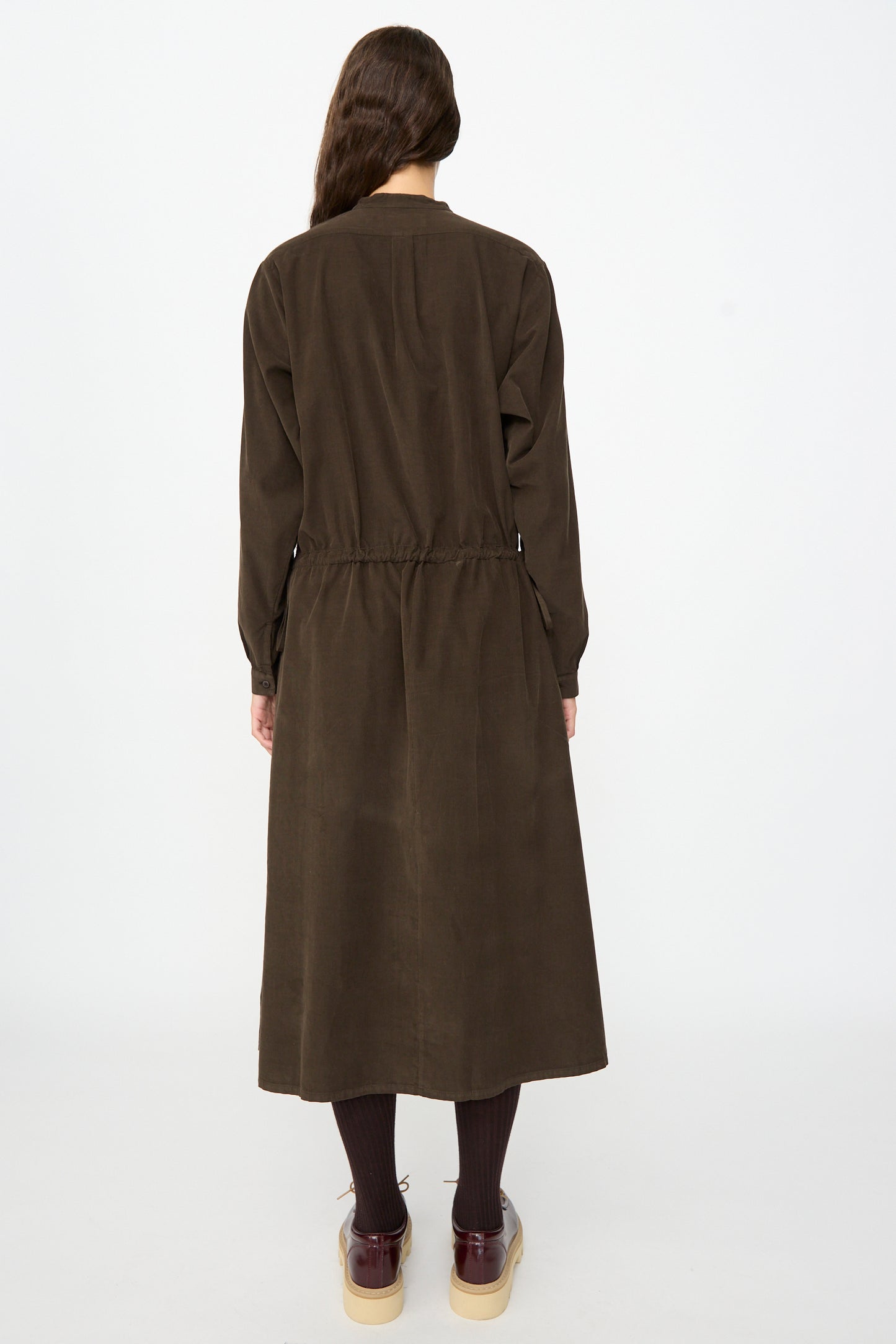 A person stands facing away, wearing the Labo.Art Clio Dress in Cacao—an ankle-length, long-sleeved cotton needlecord dress with an elastic waist—paired with dark tights and brown shoes. The dress is made in Italy. The background is plain white.