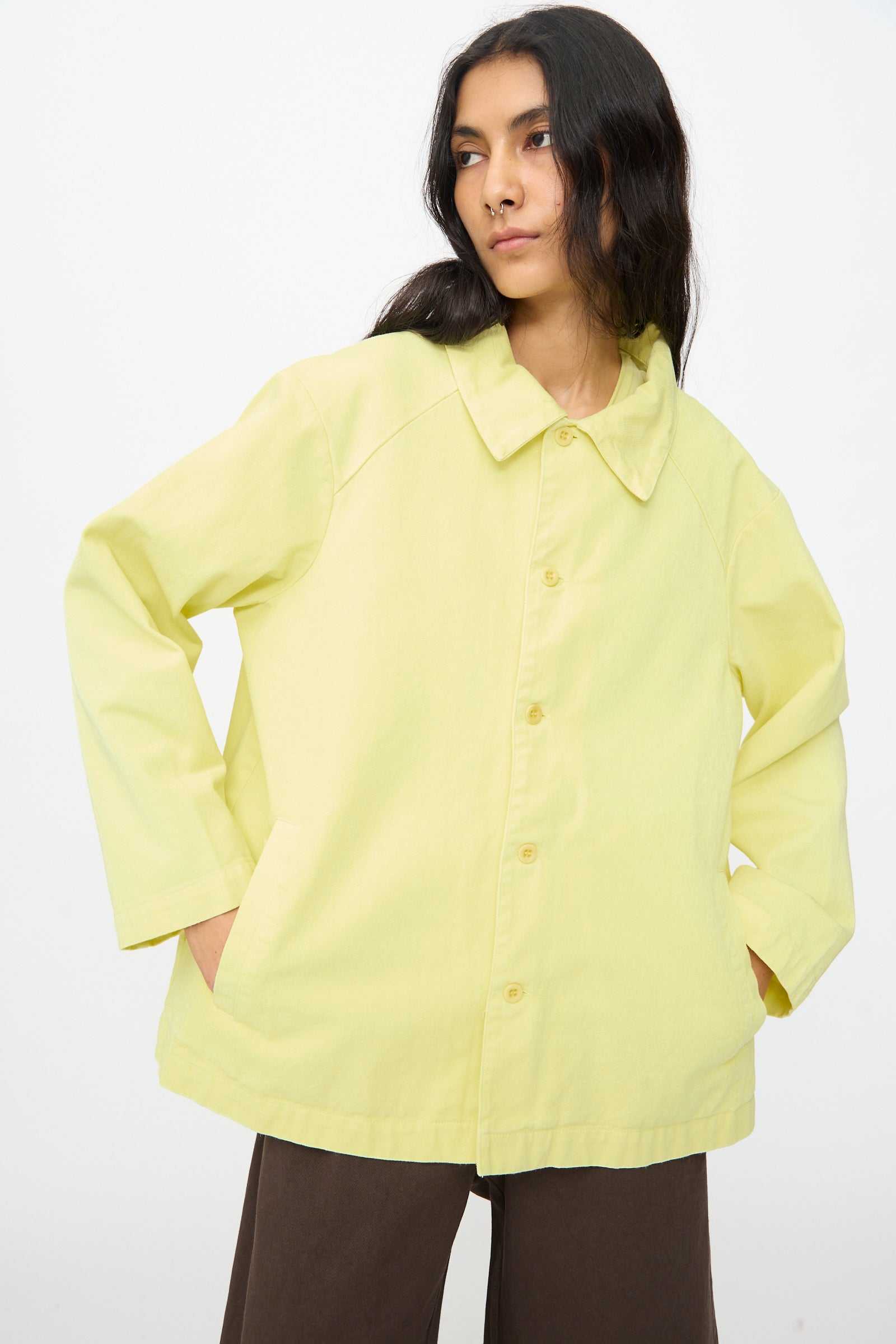 A person wears the Labo.Art Cotton Cortina Jacket in Citronella with brown pants, standing against a plain white background.