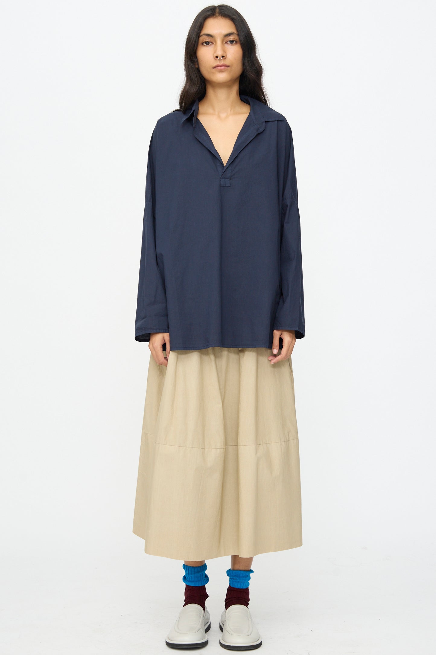 Someone stands facing forward against a plain white background wearing a loose navy long-sleeve shirt, blue and maroon socks, white slip-on shoes, and the Labo.Art Cotton Gea Skirt in Sonora.