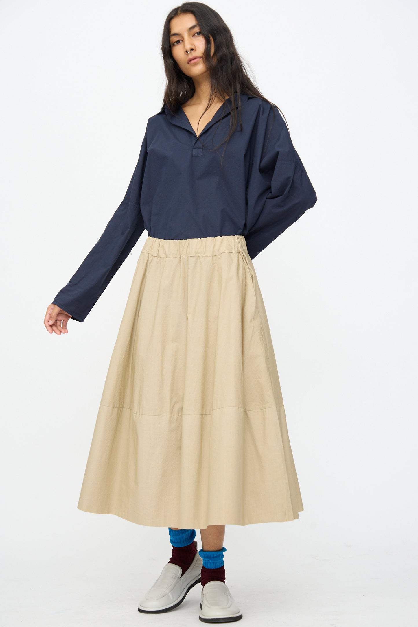 A person wears a loose navy long-sleeve shirt, Labo.Art’s Cotton Gea Skirt in Sonora, blue and maroon socks, and white slip-on shoes while standing against a plain white background.