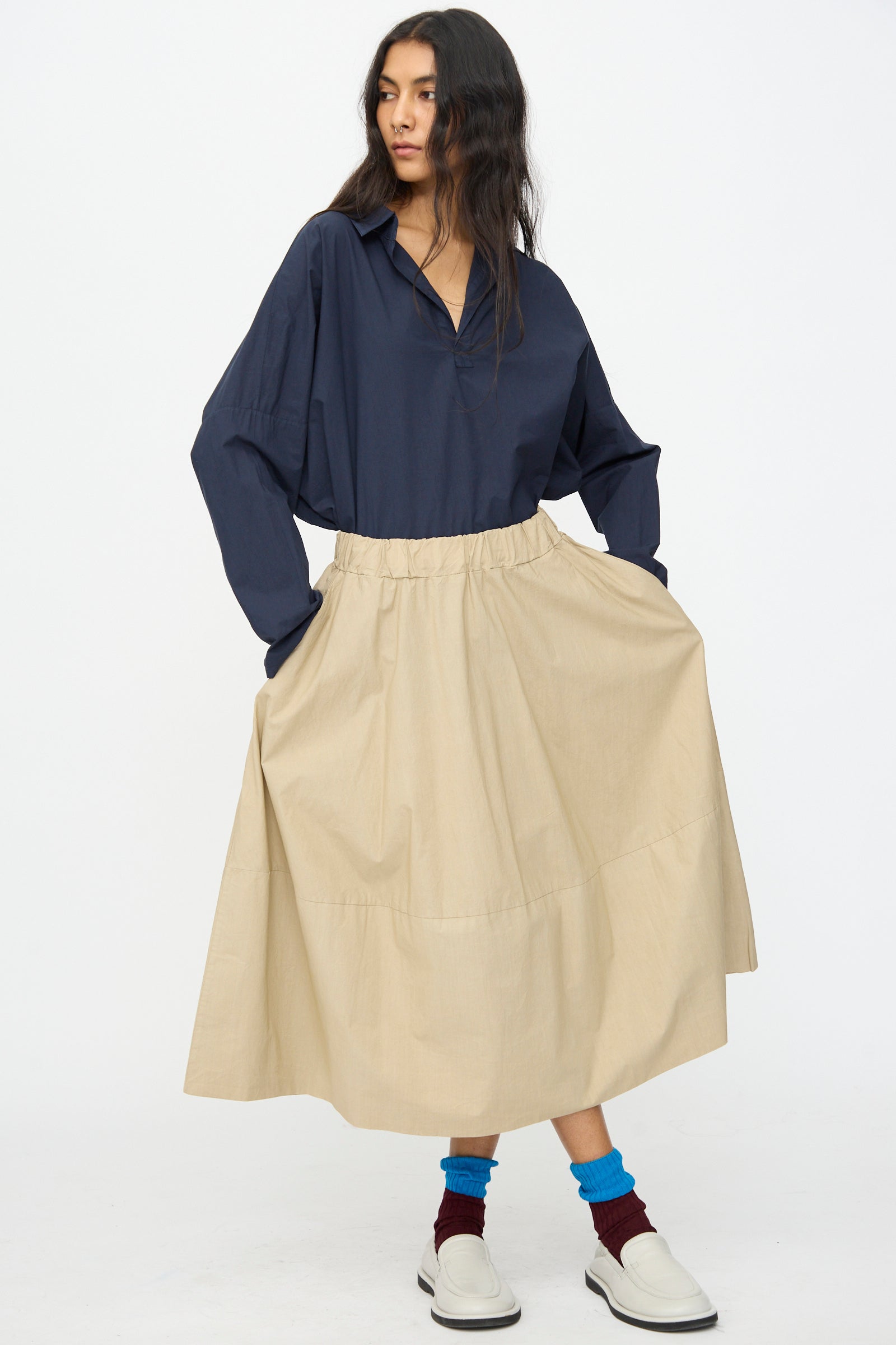 A person stands against a plain white background wearing a navy long-sleeve shirt, blue and burgundy socks, white slip-on shoes, and the Labo.Art Cotton Gea Skirt in Sonora—a cotton A-line midi skirt made in Italy.