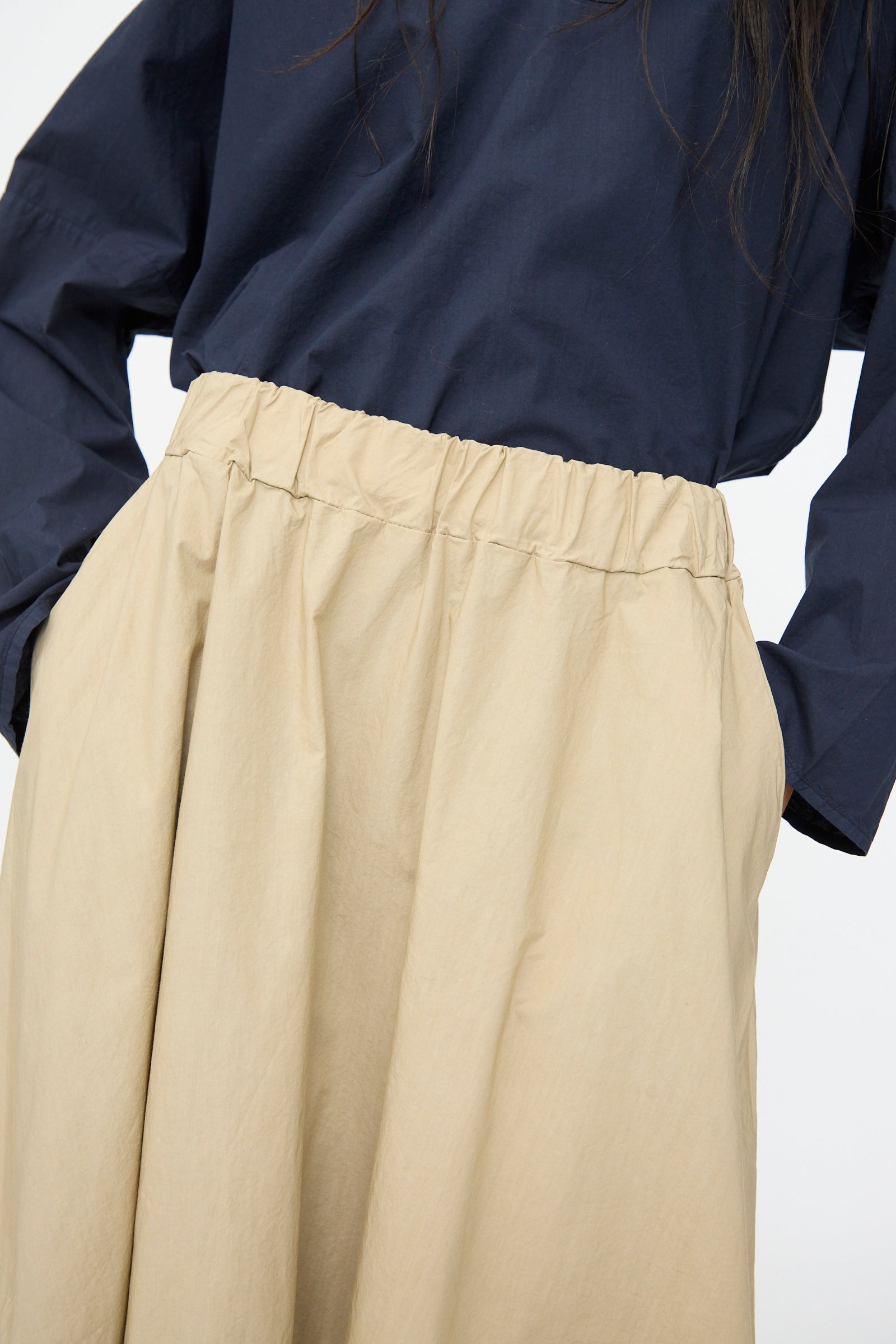 A person stands with hands in the pockets of the Labo.Art Cotton Gea Skirt in Sonora, a light beige, high-waisted midi skirt featuring an A-line silhouette and gathered details, paired with a navy long-sleeve top.