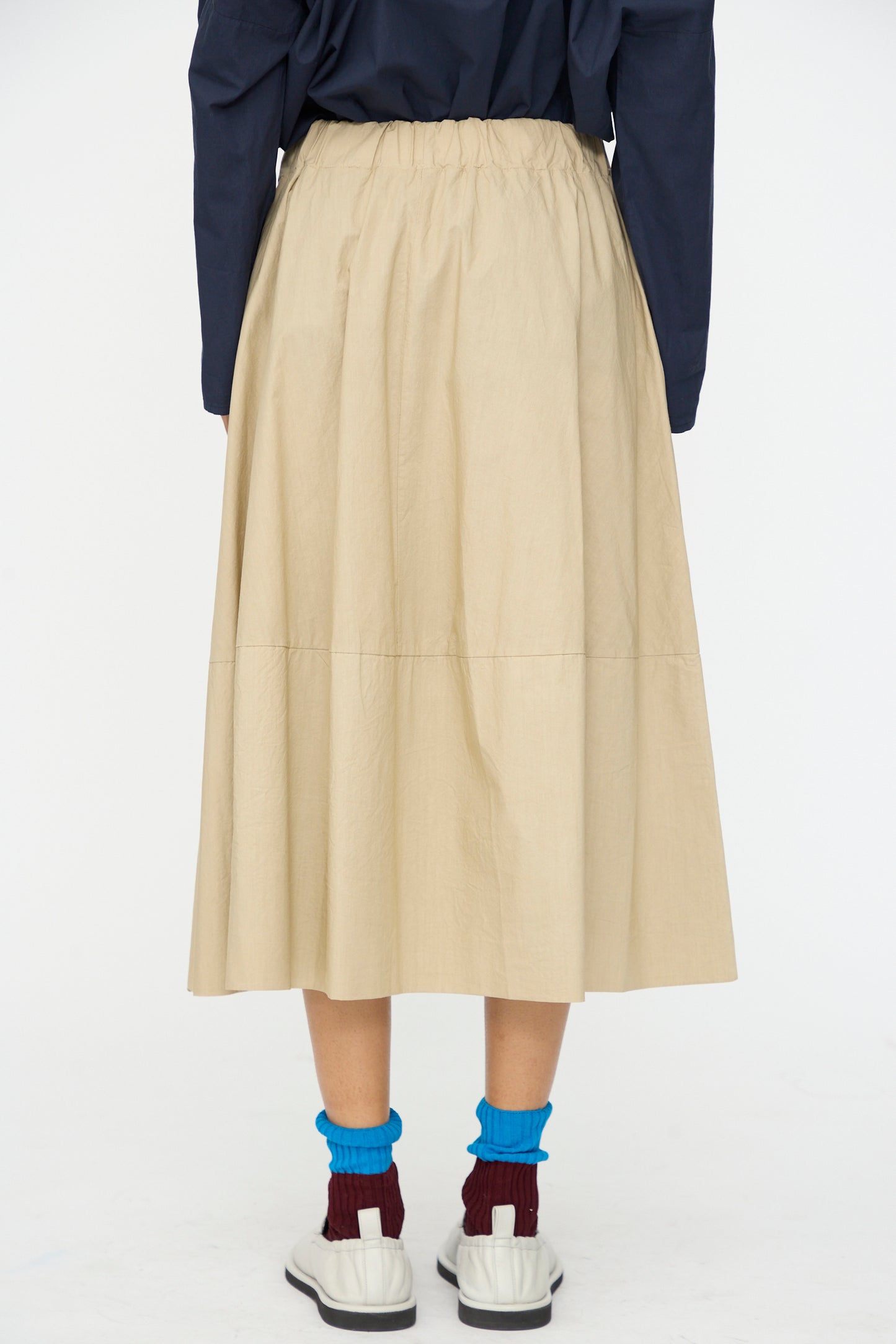 Person stands with back to camera wearing the Labo.Art Cotton Gea Skirt in Sonora, paired with a navy top, white shoes, blue socks, and burgundy socks underneath.