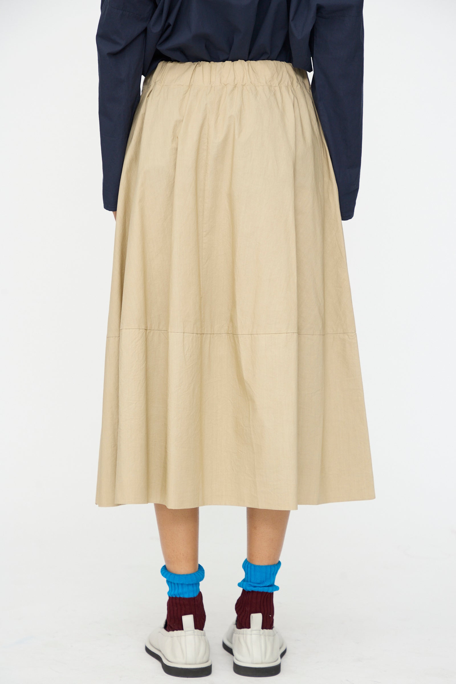 Person stands with back to camera wearing the Labo.Art Cotton Gea Skirt in Sonora, paired with a navy top, white shoes, blue socks, and burgundy socks underneath.