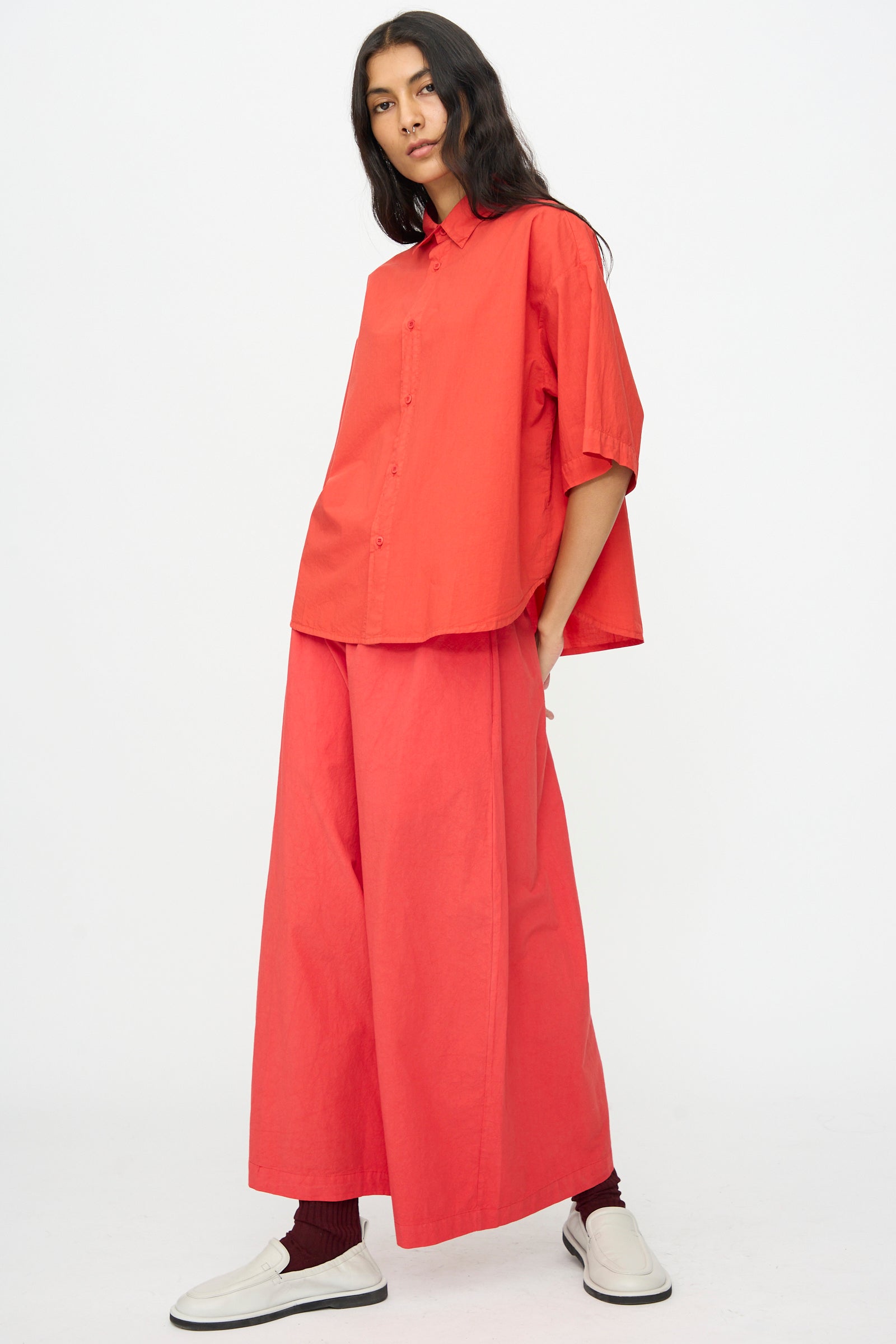 A person stands against a plain white background wearing the Labo.Art Cotton Sole Pant in Tango, matched with a loose-fitting red button-up shirt, white slip-on shoes, and dark socks.