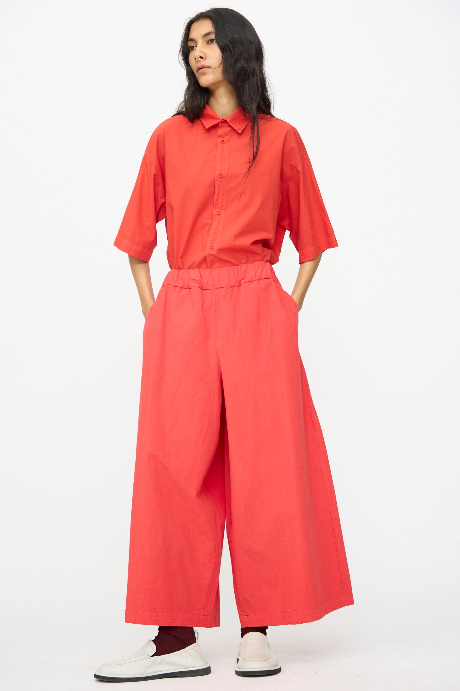 A person stands against a white background wearing a loose red button-up shirt, Labo.Art Cotton Sole Pant in Tango (wide-leg, made in Italy), red socks, and white slip-on shoes, with hands in their pockets.