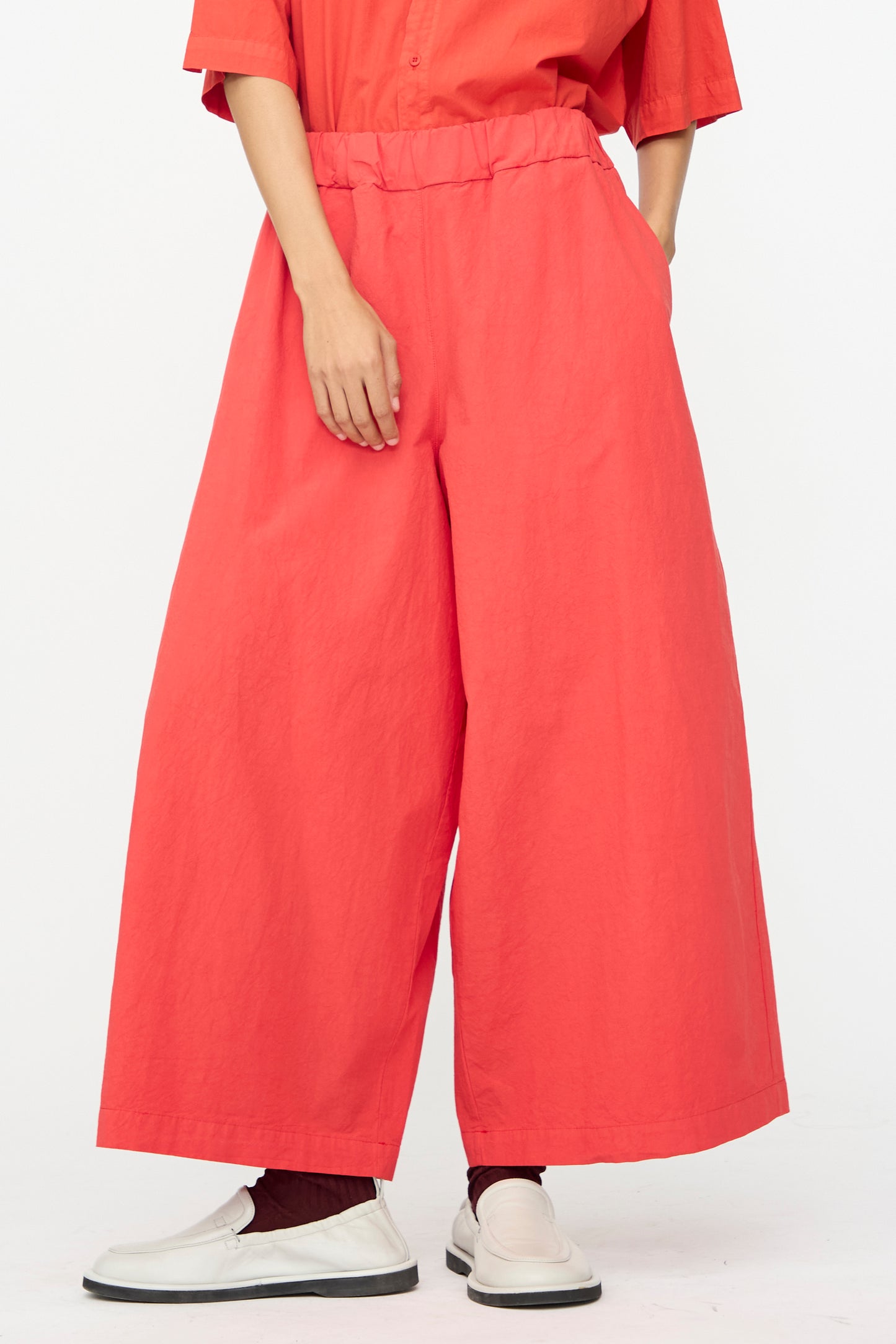 A person wears the Labo.Art Cotton Sole Pant in Tango—wide-leg, high-waisted red cotton pants made in Italy—with hands in pockets, paired with a red shirt, white slip-on shoes, and red socks against a plain white background.