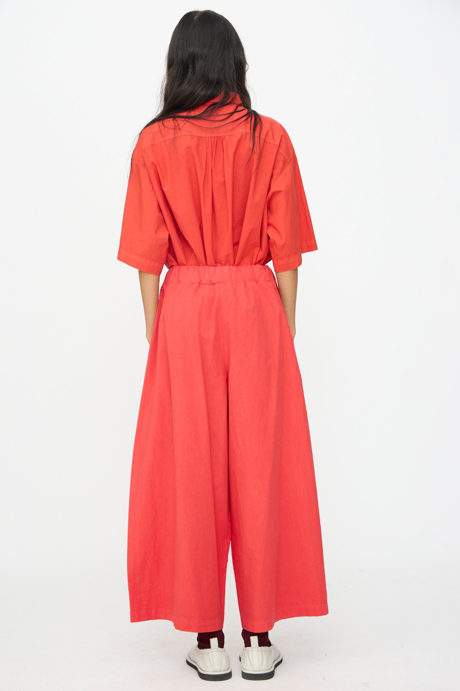 A person stands with their back to the camera, wearing the Labo.Art Cotton Sole Pant in Tango and white slip-on shoes against a plain white background.