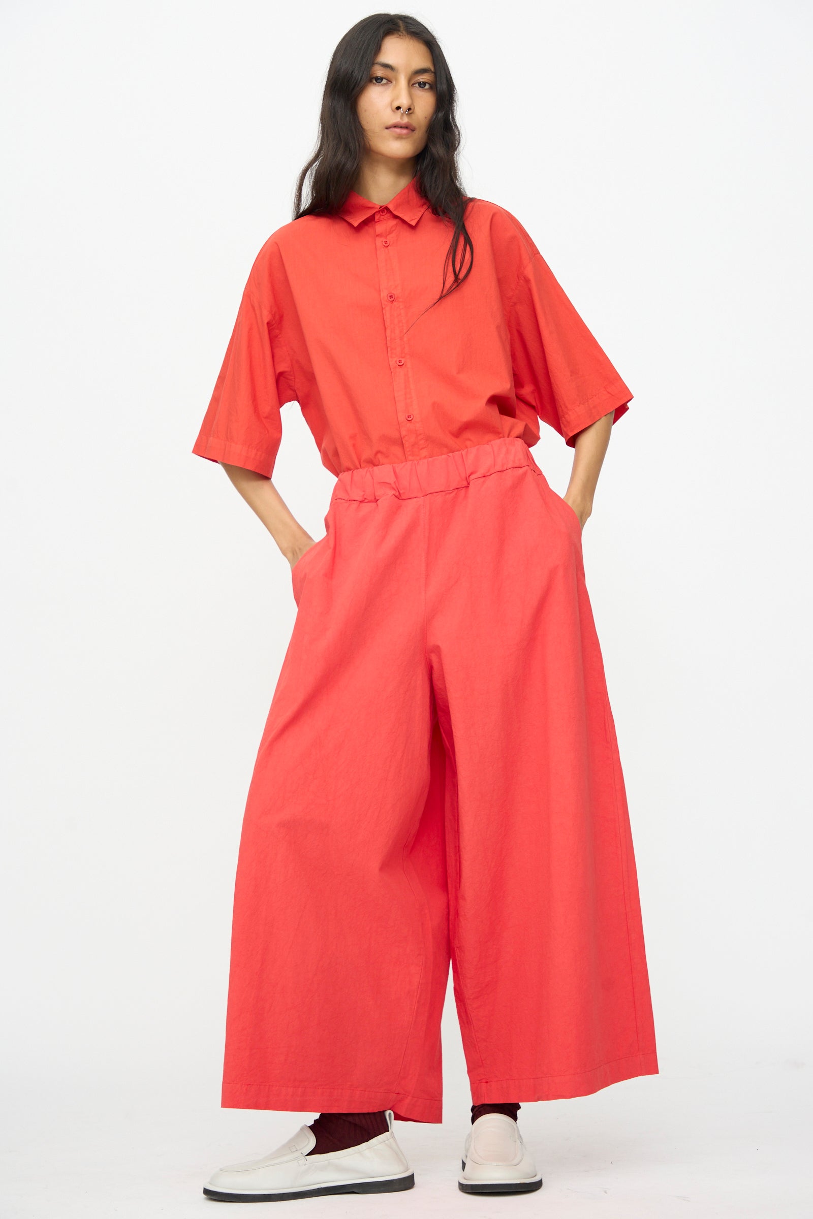 A person stands against a plain background wearing Labo.Art’s loose-fitting Cotton Sole Pant in Tango and white slip-on shoes.