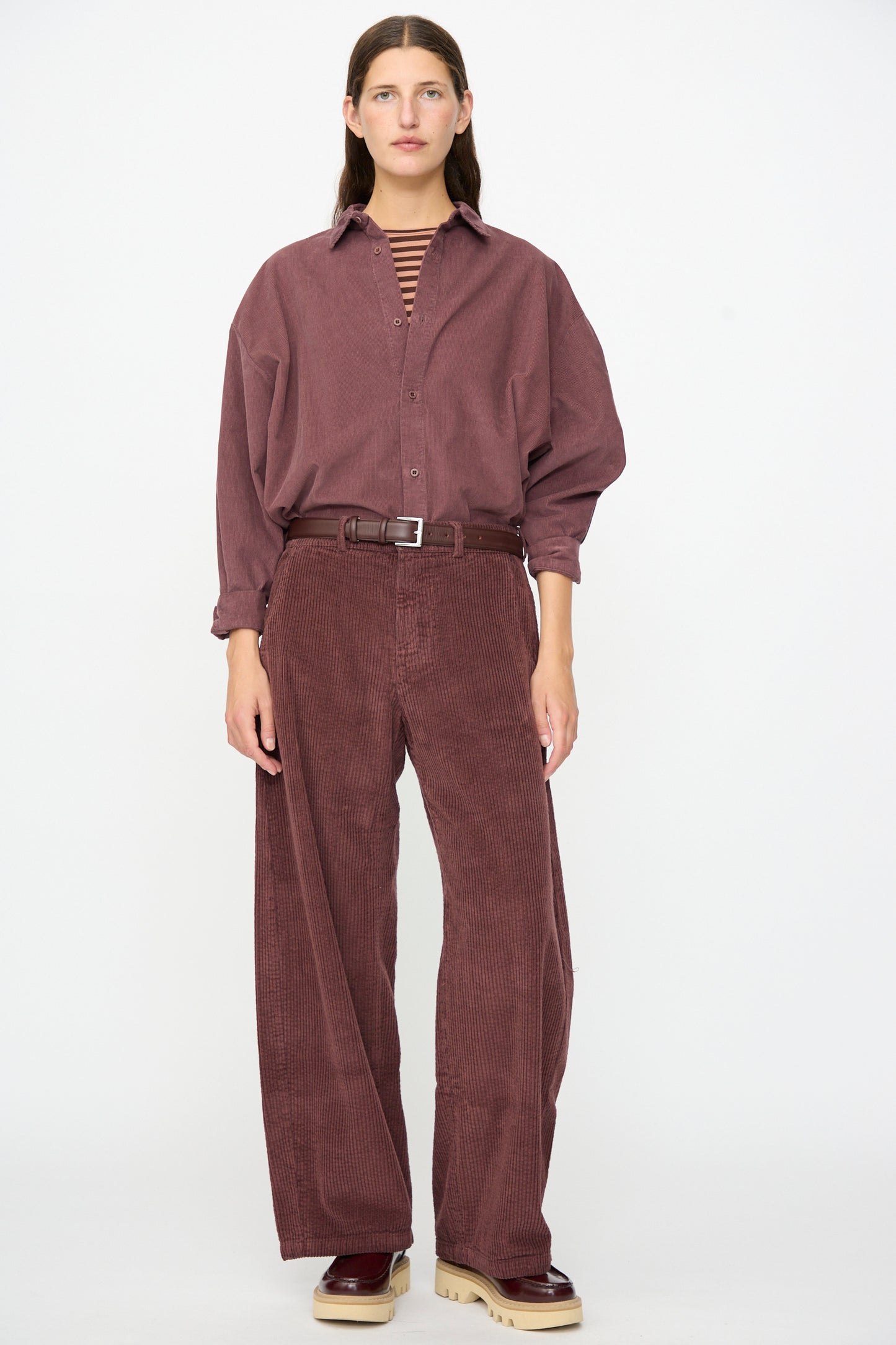 A woman stands facing forward against a plain white background, wearing Labo.Art’s Cotton Trouser in Sasso styled with a loose maroon corduroy shirt, a striped shirt underneath, a brown belt, and chunky beige-soled shoes.