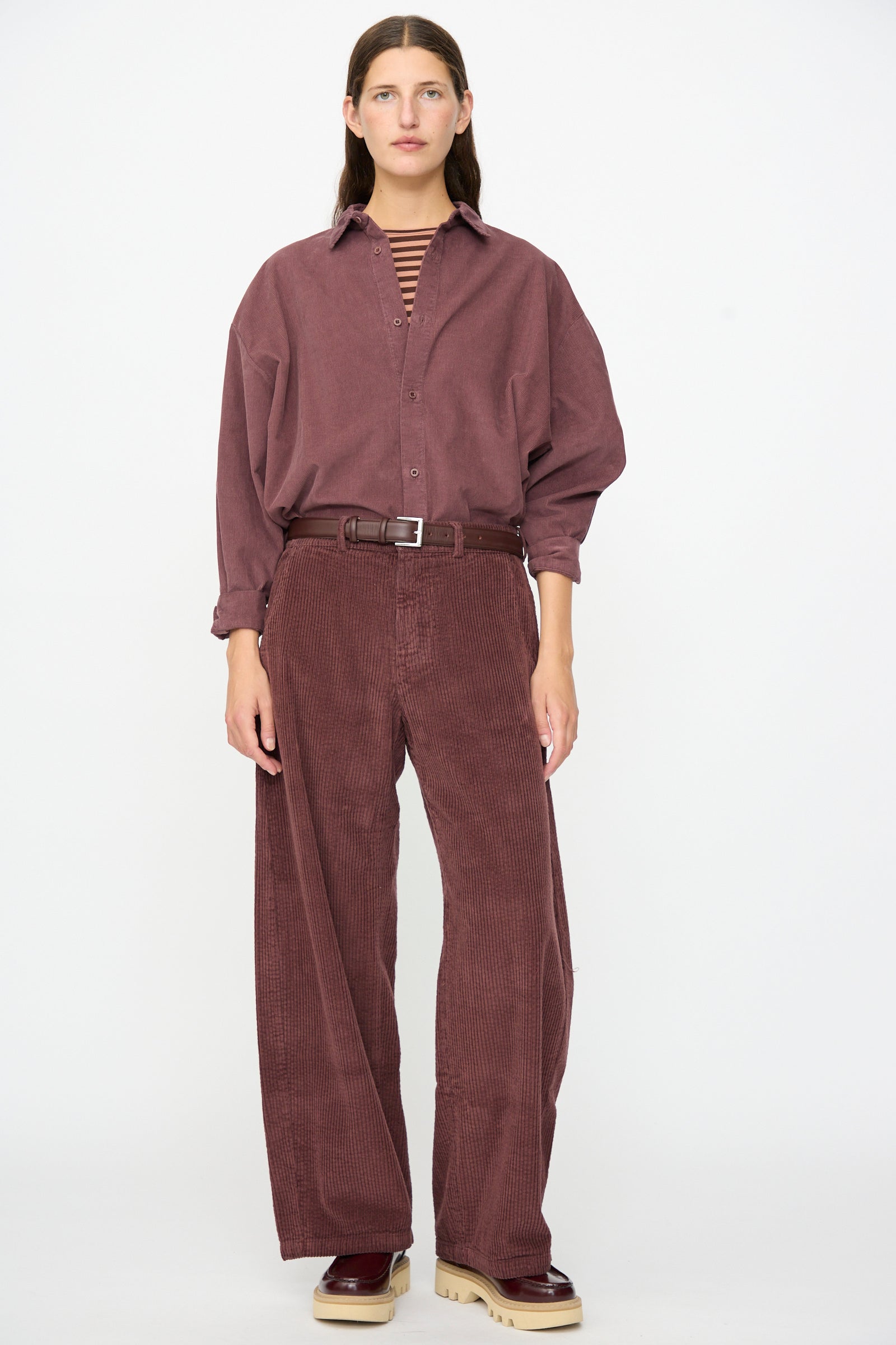A woman stands facing forward against a plain white background, wearing Labo.Art’s Cotton Trouser in Sasso styled with a loose maroon corduroy shirt, a striped shirt underneath, a brown belt, and chunky beige-soled shoes.