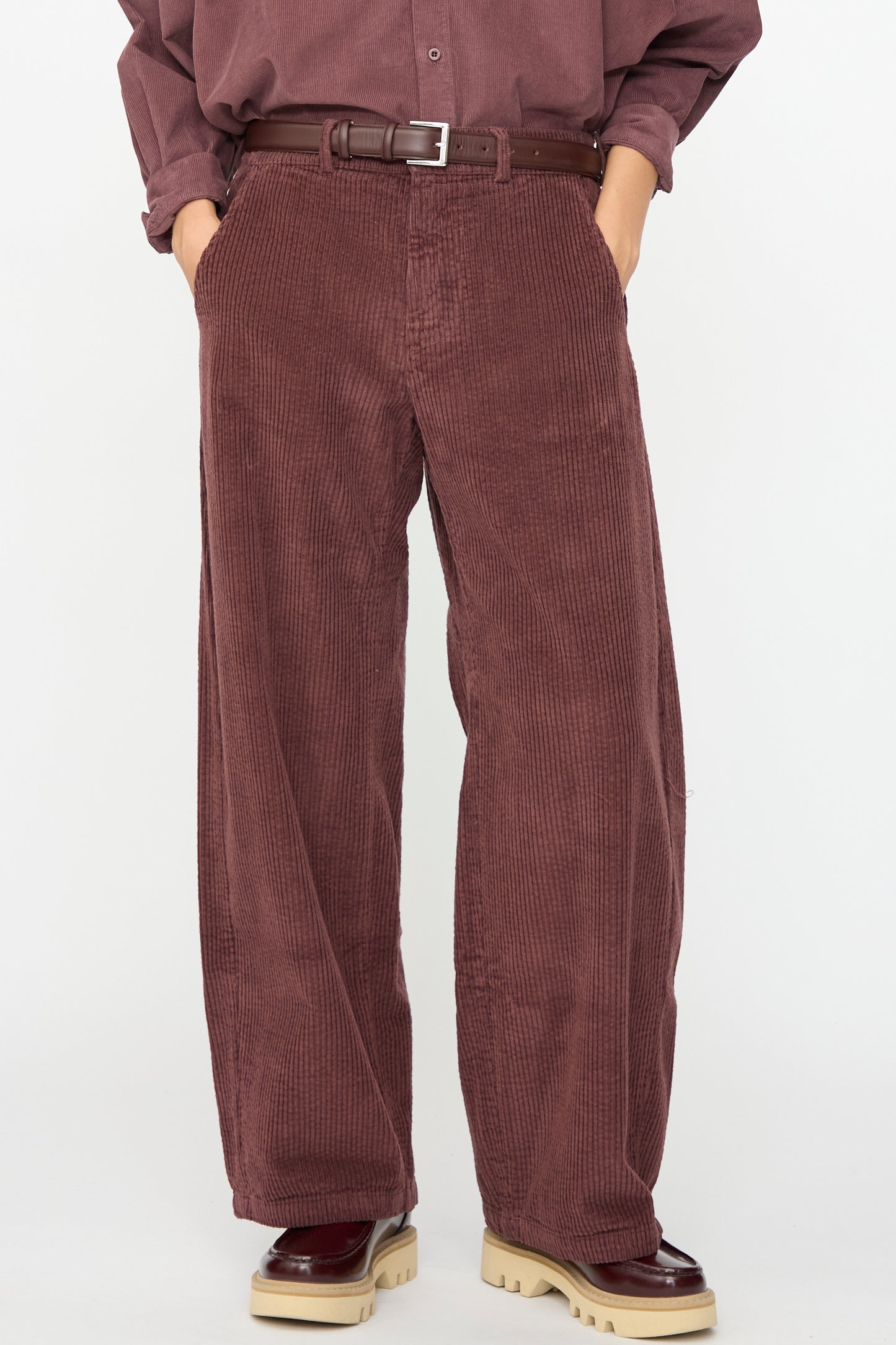 A person wearing Labo.Art’s Cotton Trouser in Sasso—wide-leg, relaxed fit corduroy pants made in Italy—paired with a brown belt, matching shirt, and two-toned shoes, stands with hands in pockets against a plain background.