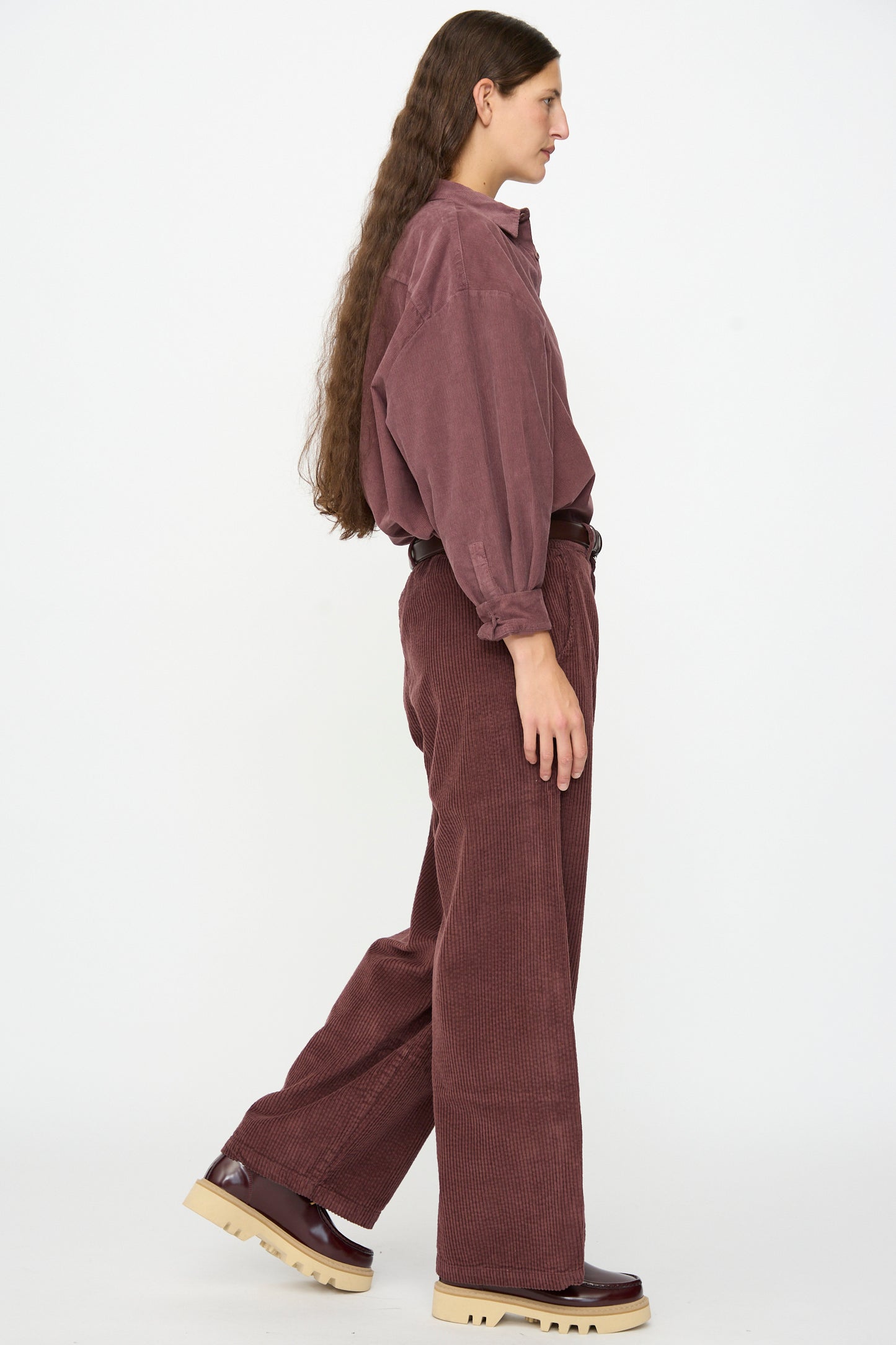 A person with long brown hair stands in profile against a plain white background, dressed in a loose maroon corduroy shirt and wide-leg Labo.Art Cotton Trouser in Sasso, made in Italy. The look is finished with a brown belt and chunky brown shoes.