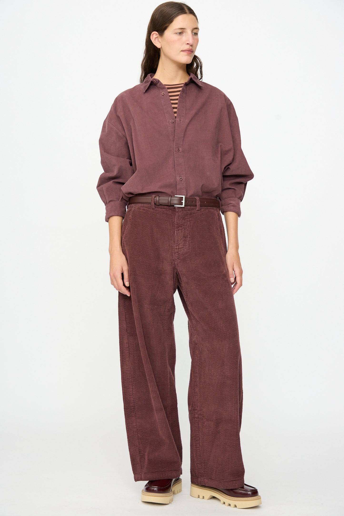A person wearing the Labo.Art Cotton Trouser in Sasso, featuring a relaxed fit silhouette, styled with a loose maroon corduroy shirt, a striped top layered underneath, and beige-soled shoes, stands against a plain white background.