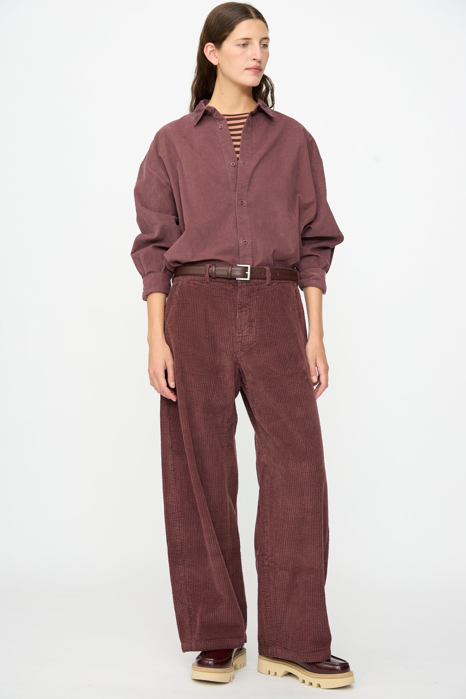 A person wearing the Labo.Art Cotton Trouser in Sasso, featuring a relaxed fit silhouette, styled with a loose maroon corduroy shirt, a striped top layered underneath, and beige-soled shoes, stands against a plain white background.