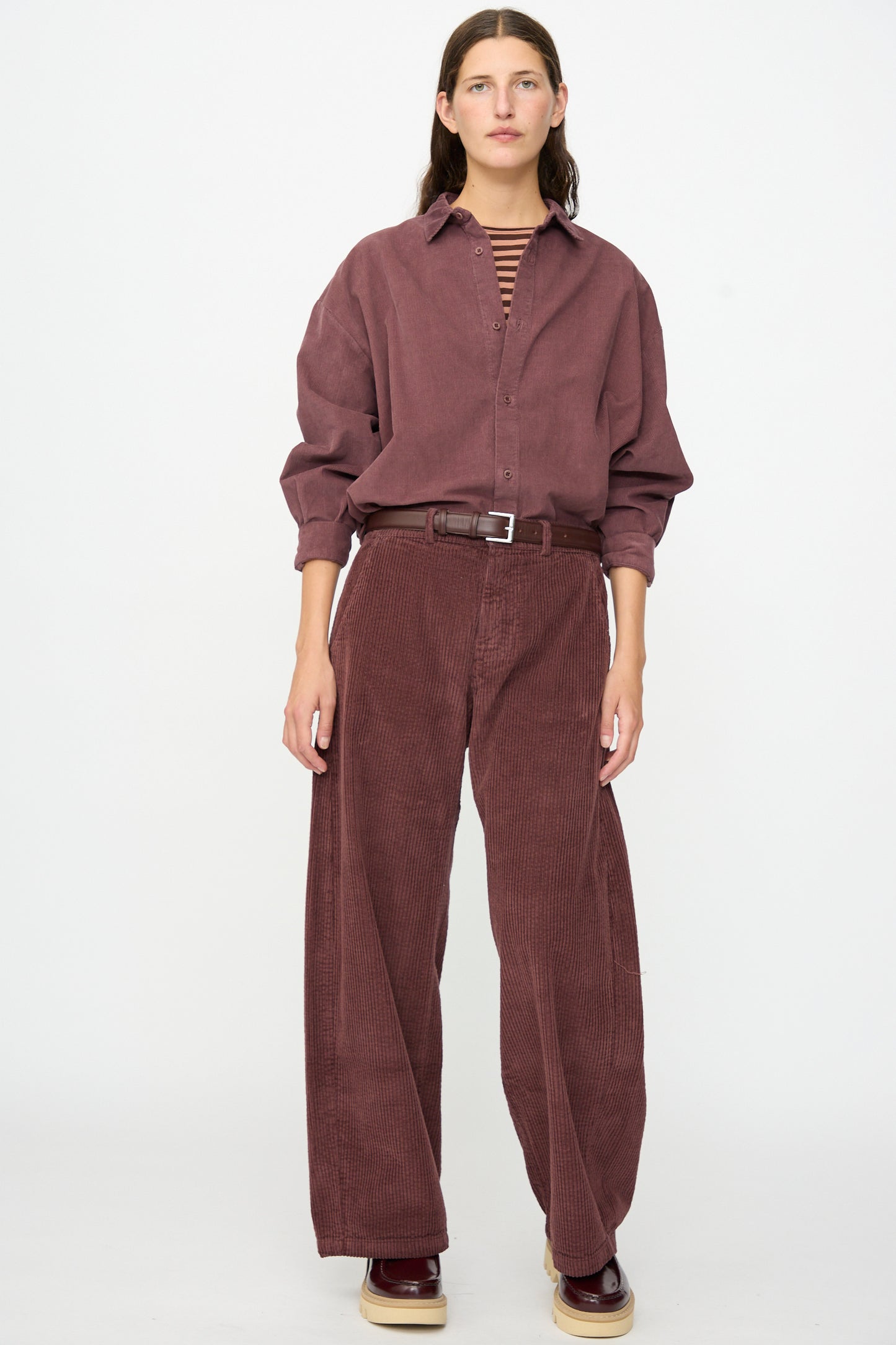 A person stands facing forward, wearing a loose burgundy corduroy shirt paired with Labo.Art’s Cotton Trouser in Sasso, which features a relaxed fit pant silhouette, completed with a brown belt and burgundy shoes.