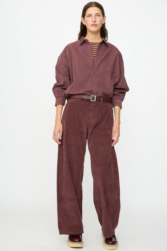 A person stands facing forward, wearing a loose burgundy corduroy shirt paired with Labo.Art’s Cotton Trouser in Sasso, which features a relaxed fit pant silhouette, completed with a brown belt and burgundy shoes.