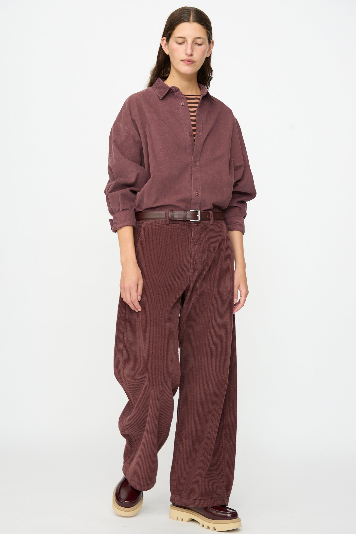 A person wearing the Labo.Art Cotton Trouser in Sasso paired with a loose burgundy corduroy shirt, a striped top underneath, a brown belt, and burgundy shoes, standing against a plain white background.