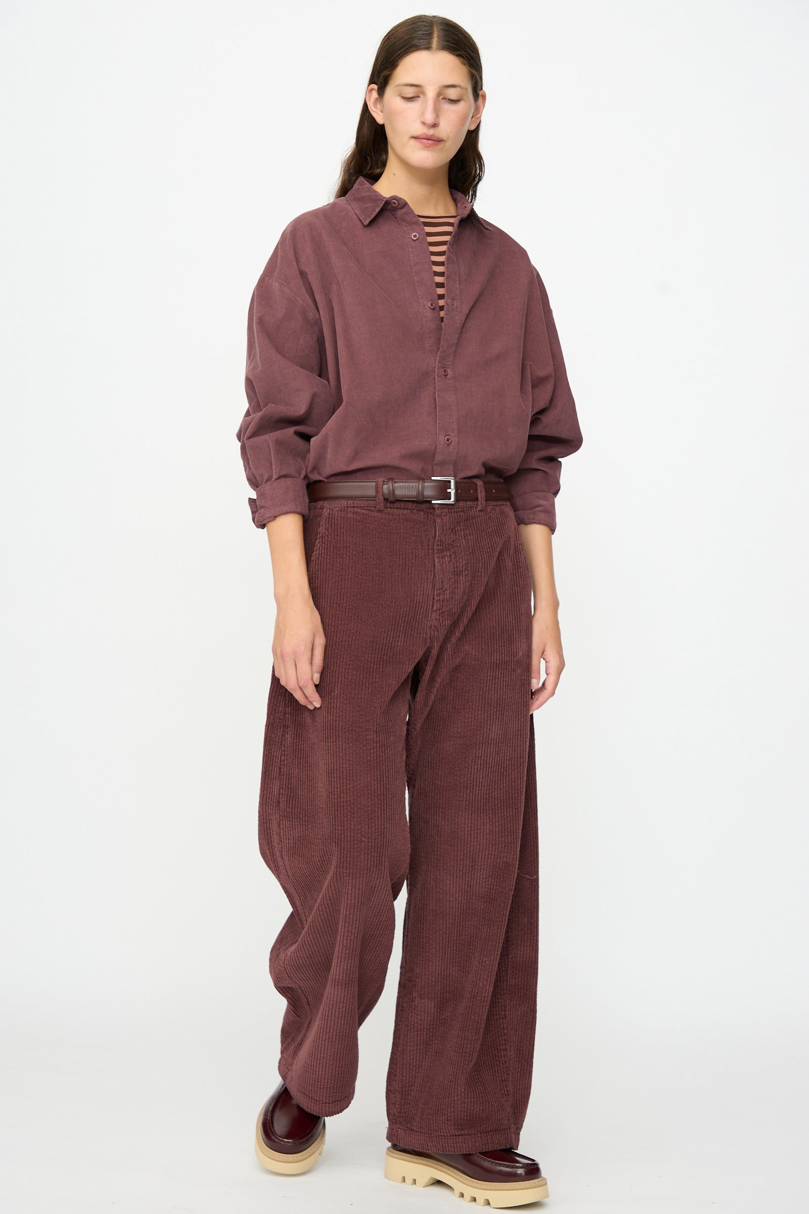 A person wearing the Labo.Art Cotton Trouser in Sasso paired with a loose burgundy corduroy shirt, a striped top underneath, a brown belt, and burgundy shoes, standing against a plain white background.