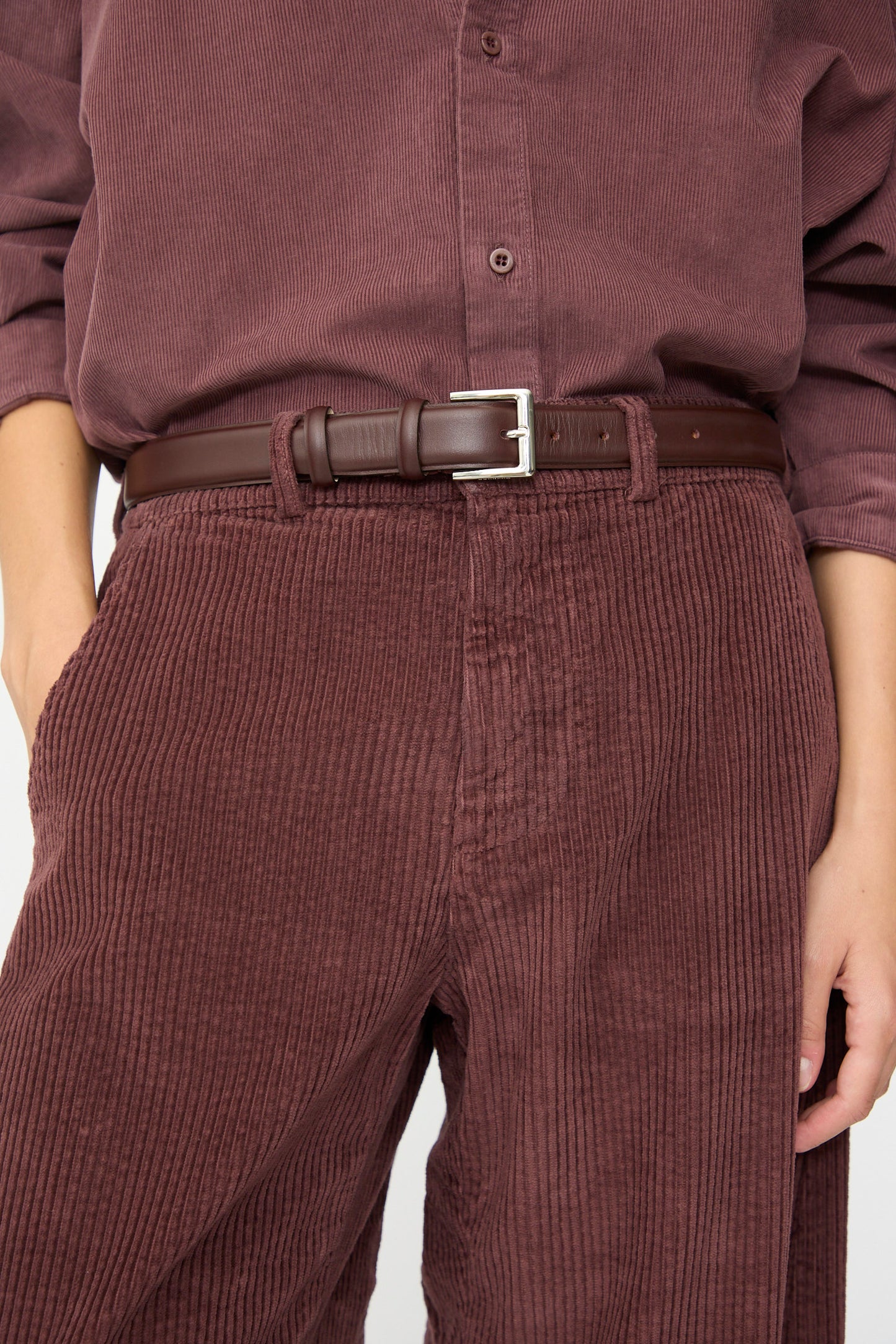 A person wearing a maroon corduroy shirt paired with Labo.Art’s Cotton Trouser in Sasso, cinched with a brown leather belt, one hand in pocket, shown from shoulder to thigh.