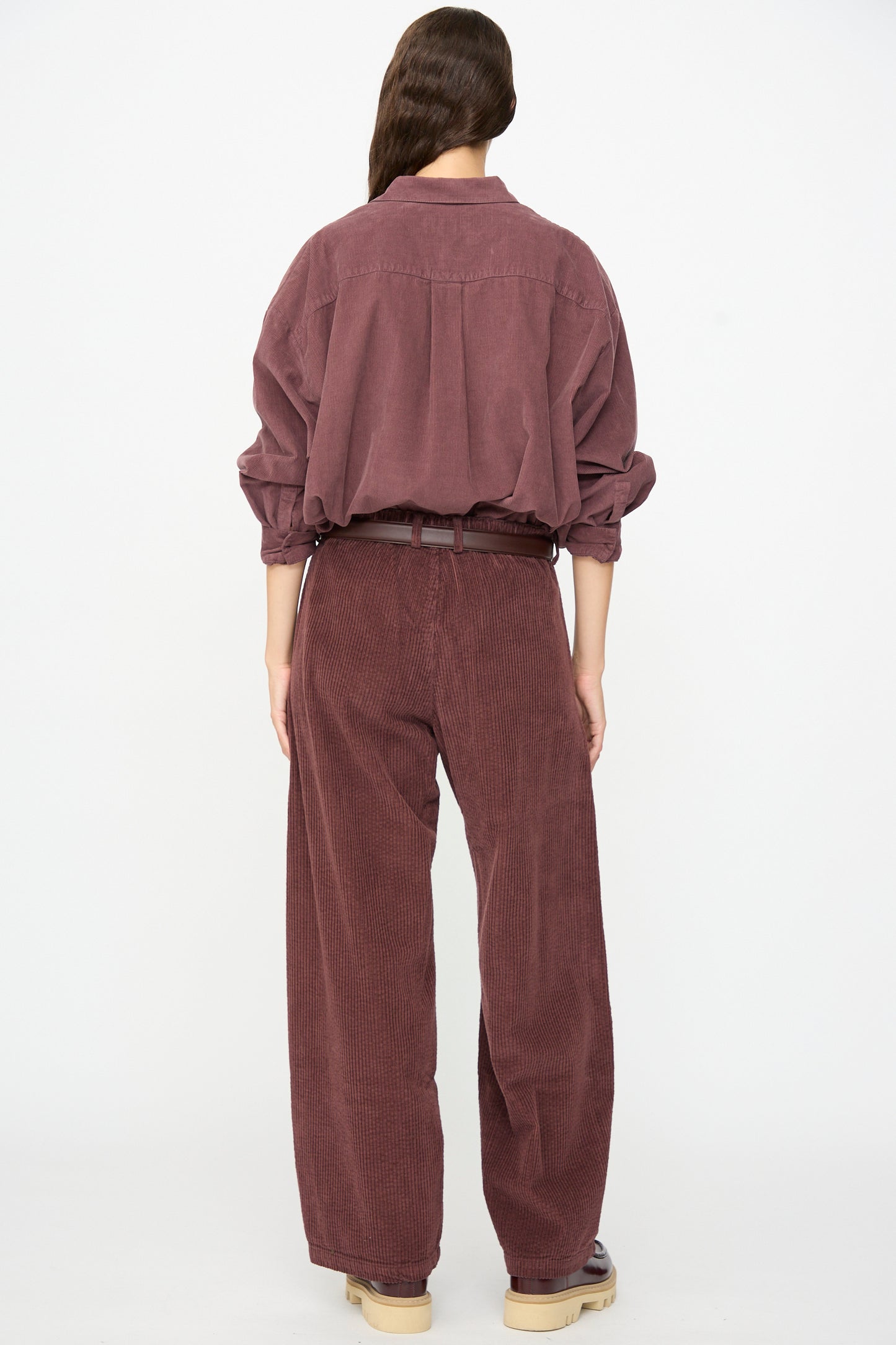 A person stands with their back to the camera, wearing a loose-fitting, long-sleeved maroon shirt paired with the Labo.Art Cotton Trouser in Sasso—a relaxed-fit style featuring a plum corduroy texture and belted waist for a chic, modern look.