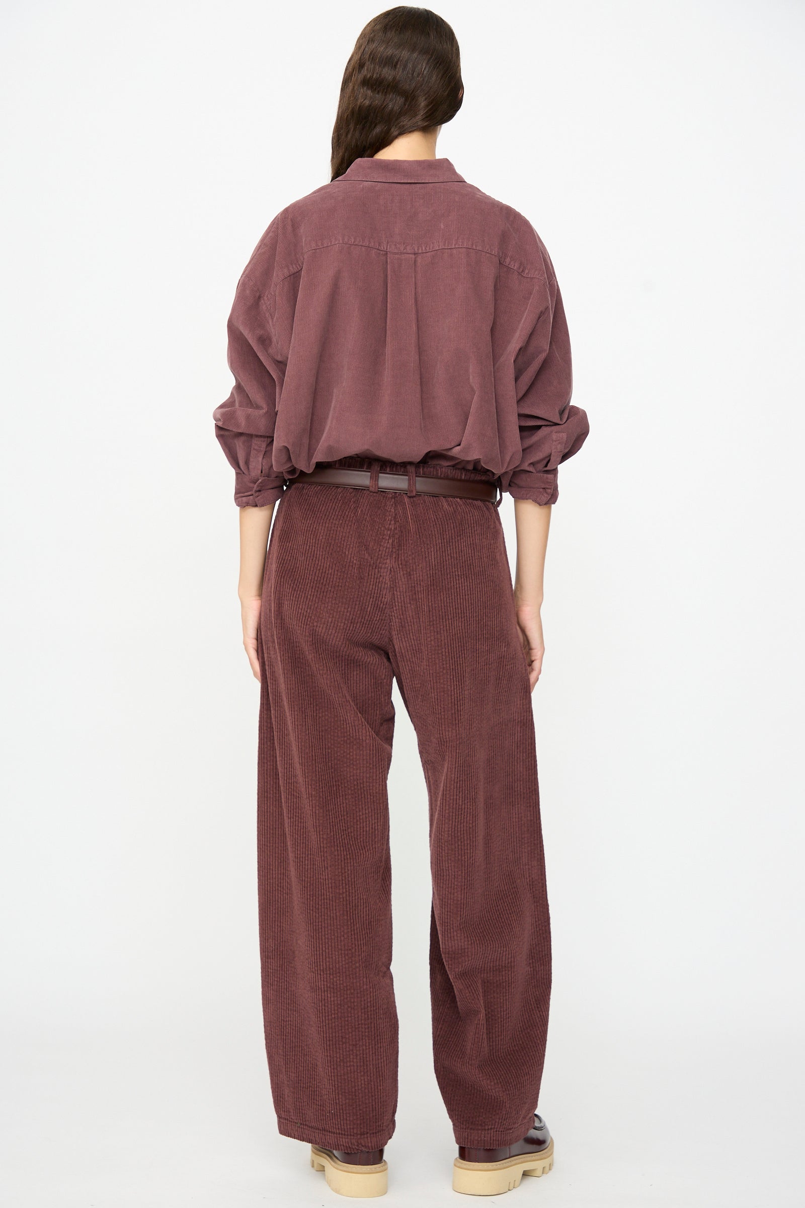 A person stands with their back to the camera, wearing a loose-fitting, long-sleeved maroon shirt paired with the Labo.Art Cotton Trouser in Sasso—a relaxed-fit style featuring a plum corduroy texture and belted waist for a chic, modern look.