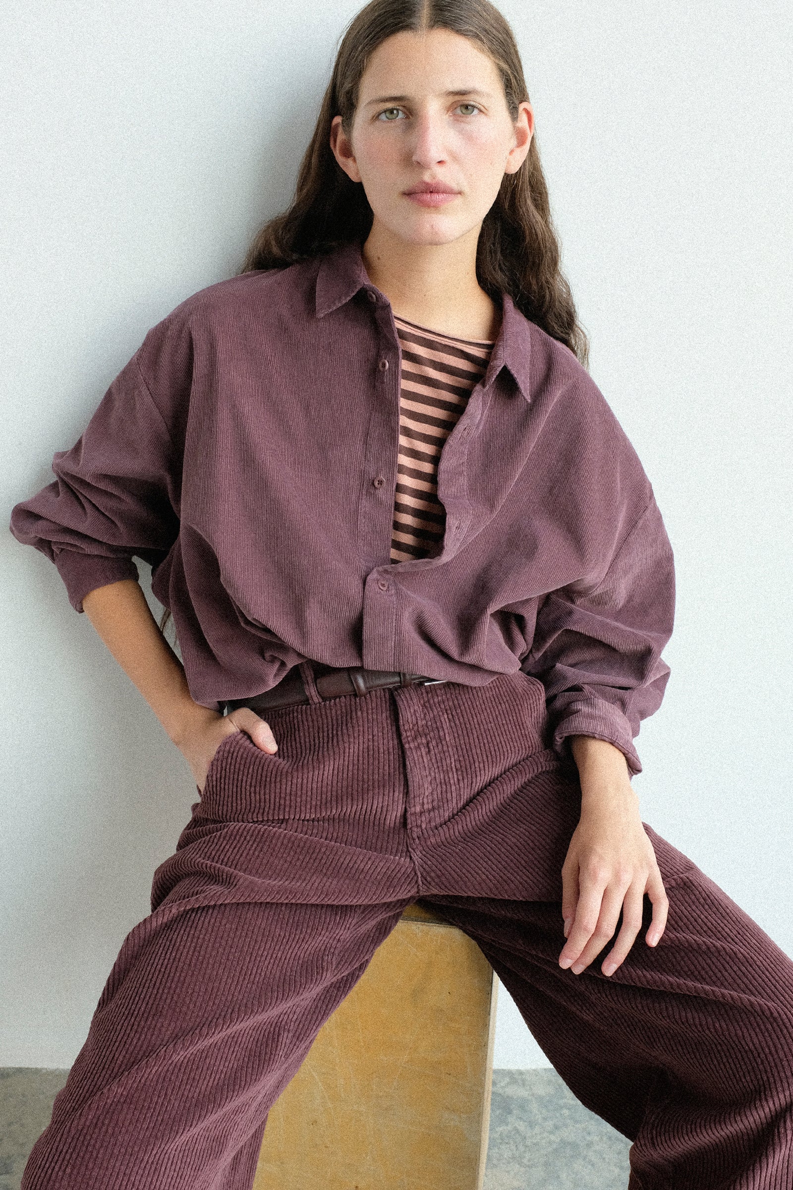 A woman Labo.Art's Quadra Shirt in Sasso with the Cotton Trouser in Sasso, sitting on a wooden block against a light gray wall.