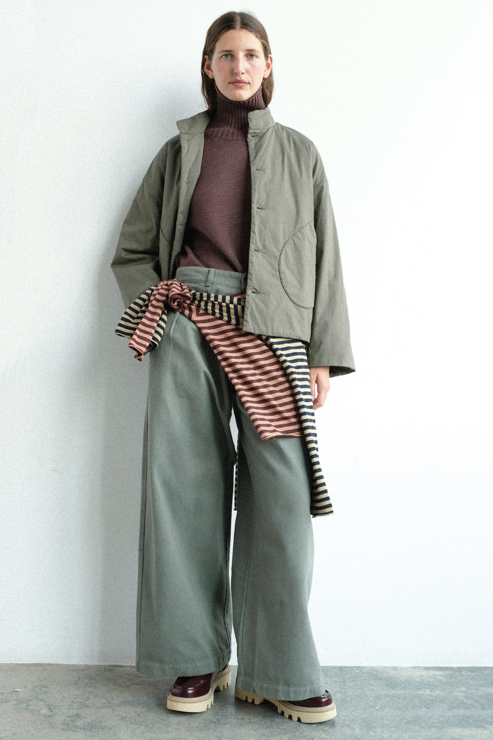 A model stands against a plain white wall wearing Labo.Art's Fiore Sweater in Sasso, Ficus Jacket in Lichene, and Marlon Trouser in Lichene, with their striped jersey tops around waist.