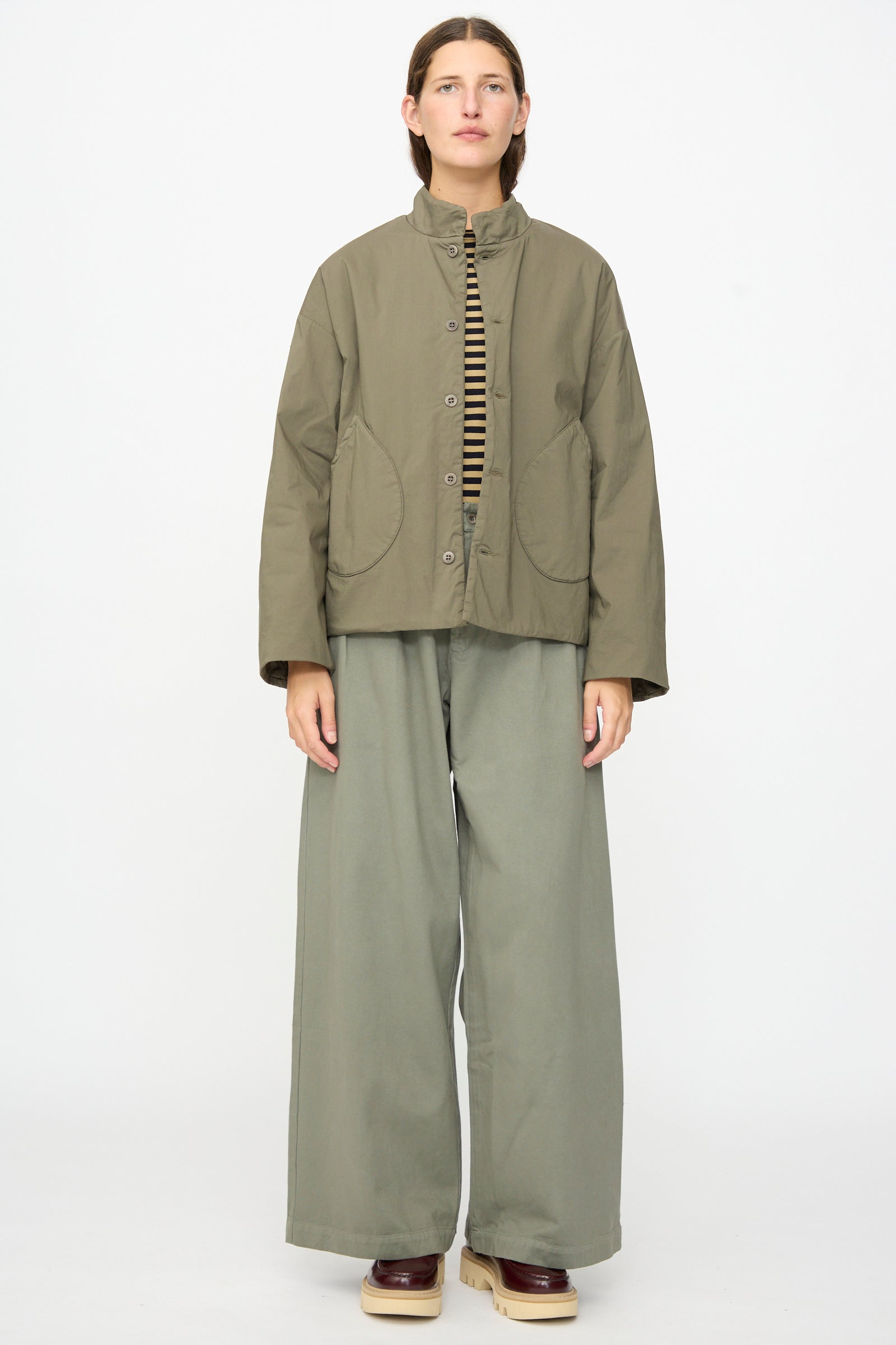 A person stands against a plain background wearing the Labo.Art Ficus Jacket in Lichene, paired with wide-leg khaki pants, a striped soft cotton-blend shirt, and brown platform shoes.