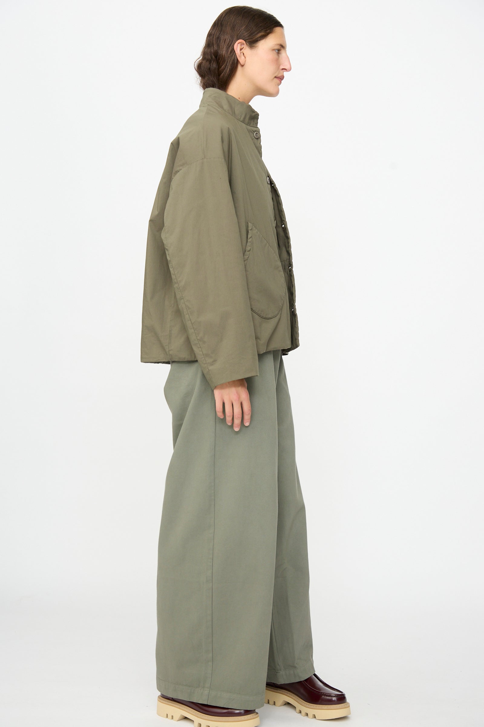 A person stands in profile against a plain white background wearing the Labo.Art Ficus Jacket in Lichene, styled with wide-leg pants and maroon platform shoes.
