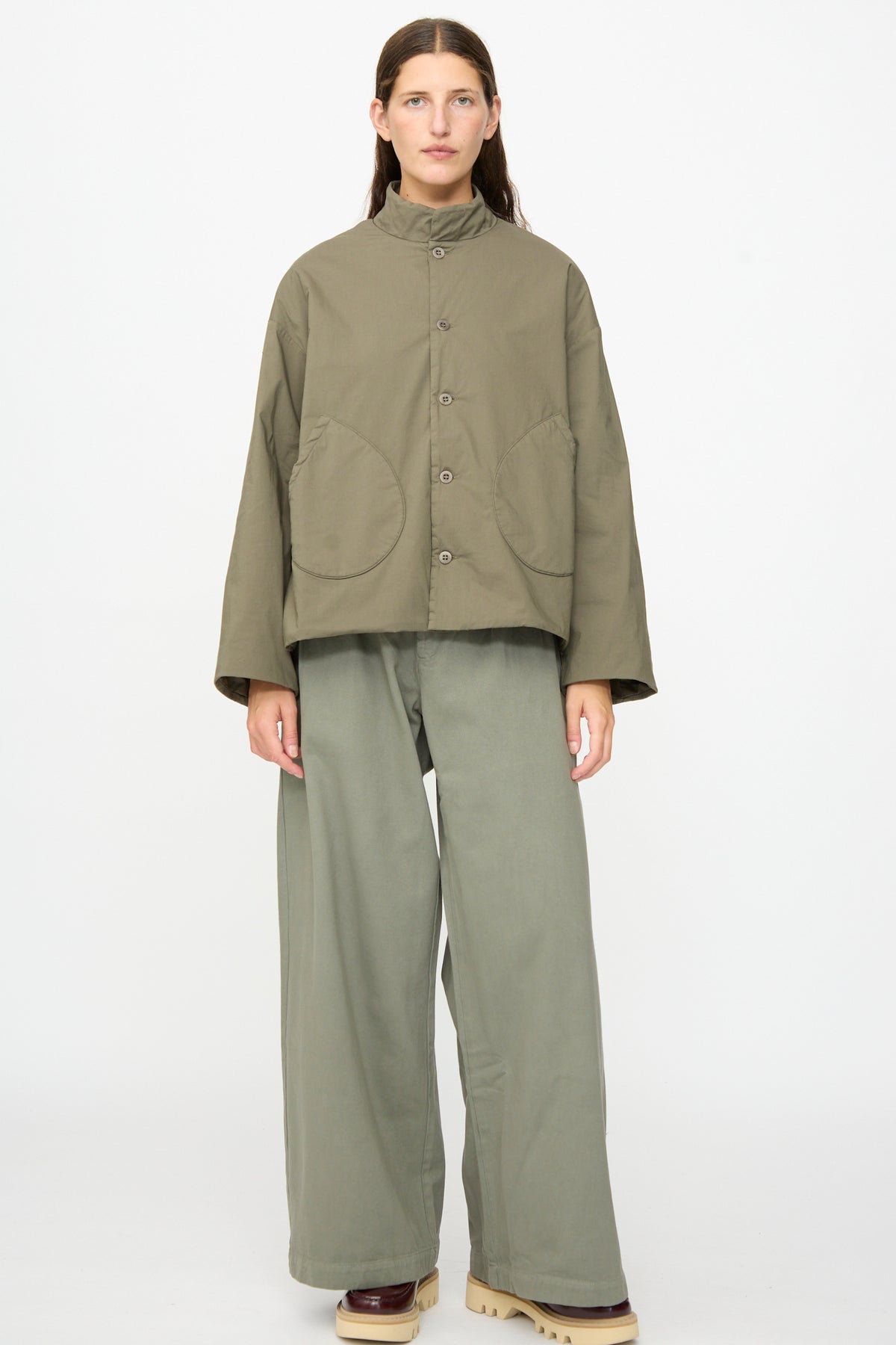 A person stands against a plain background, wearing the Labo.Art Ficus Jacket in Lichene—a loose-fitting olive green cotton blend buttoned jacket—paired with matching wide-leg pants and beige platform shoes.