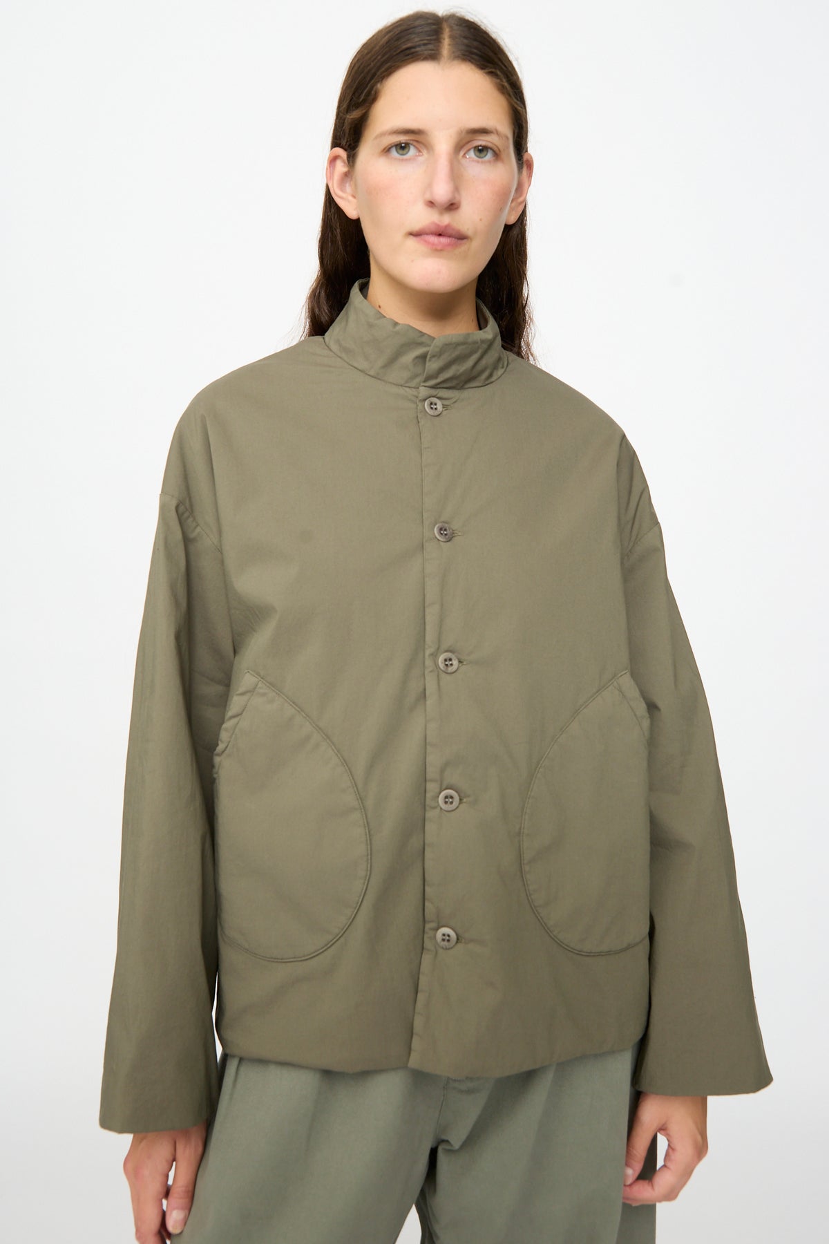 A person wearing the Labo.Art Ficus Jacket in Lichene—an olive-green, water-repellent button-up style with large front pockets and a high collar—stands against a plain light background.