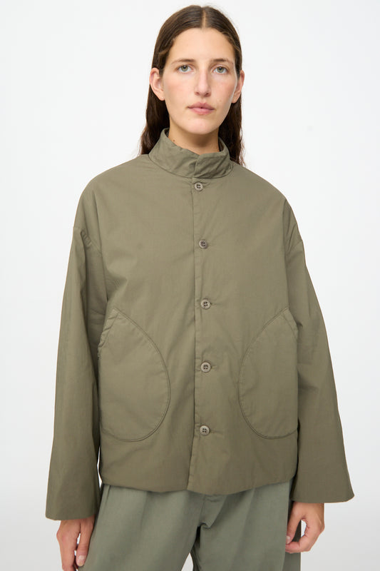 A person wearing the Labo.Art Ficus Jacket in Lichene—an olive-green, water-repellent button-up style with large front pockets and a high collar—stands against a plain light background.