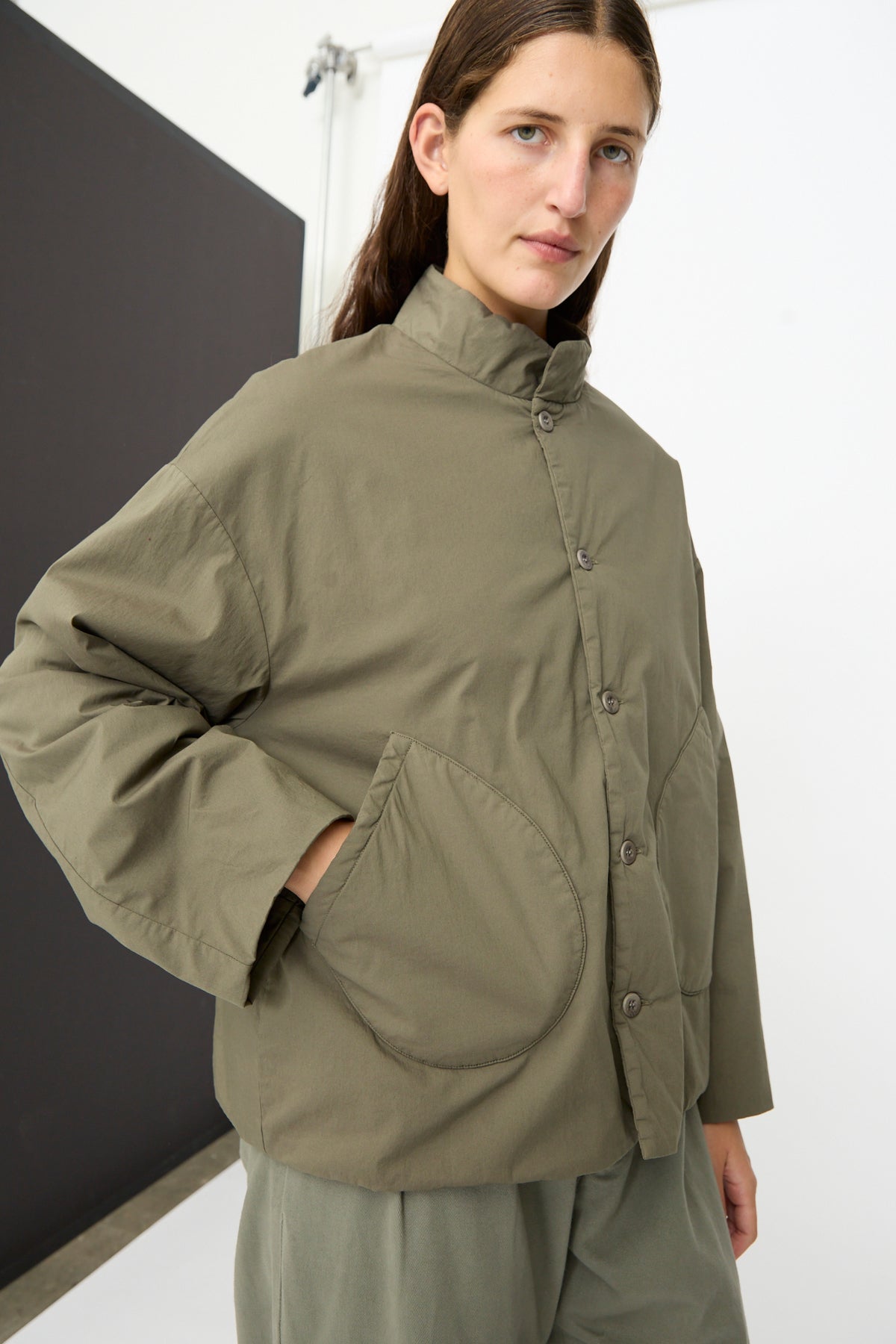 A person stands in a minimalist studio setting, wearing the Labo.Art Ficus Jacket in Lichene—an olive green, button-up padded jacket made from a water repellent cotton blend and finished with large pockets.