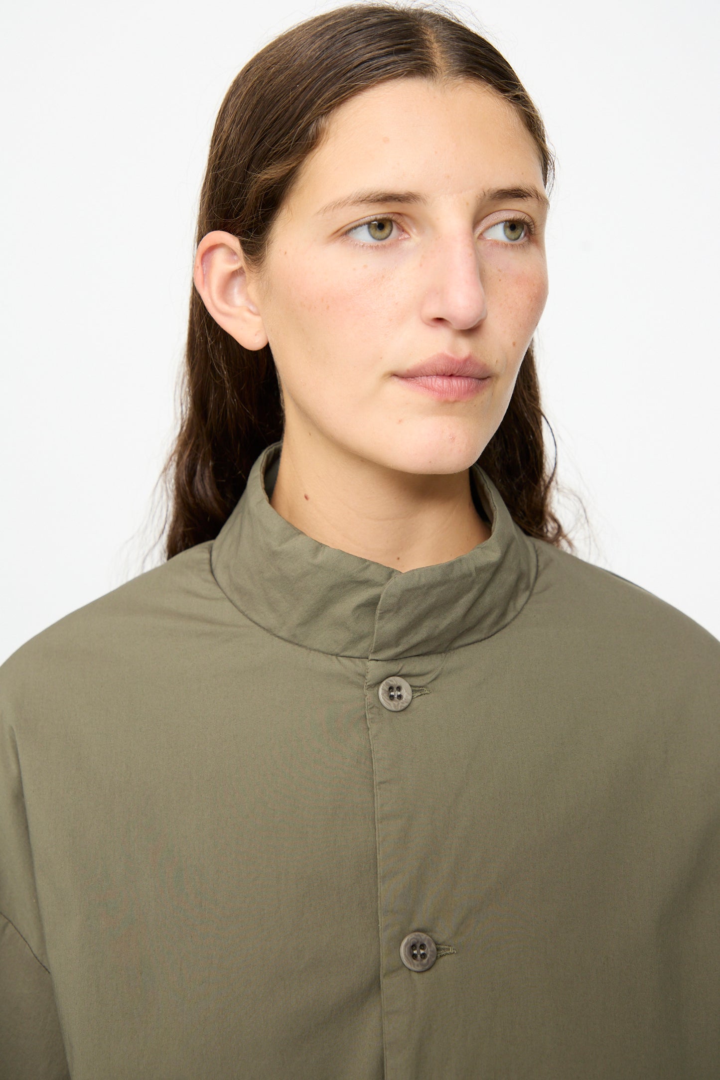 A person with long brown hair, wearing the Labo.Art Ficus Jacket in Lichene—an olive green, button-up collared padded jacket crafted from a cotton blend—looks slightly to the side against a plain white background.
