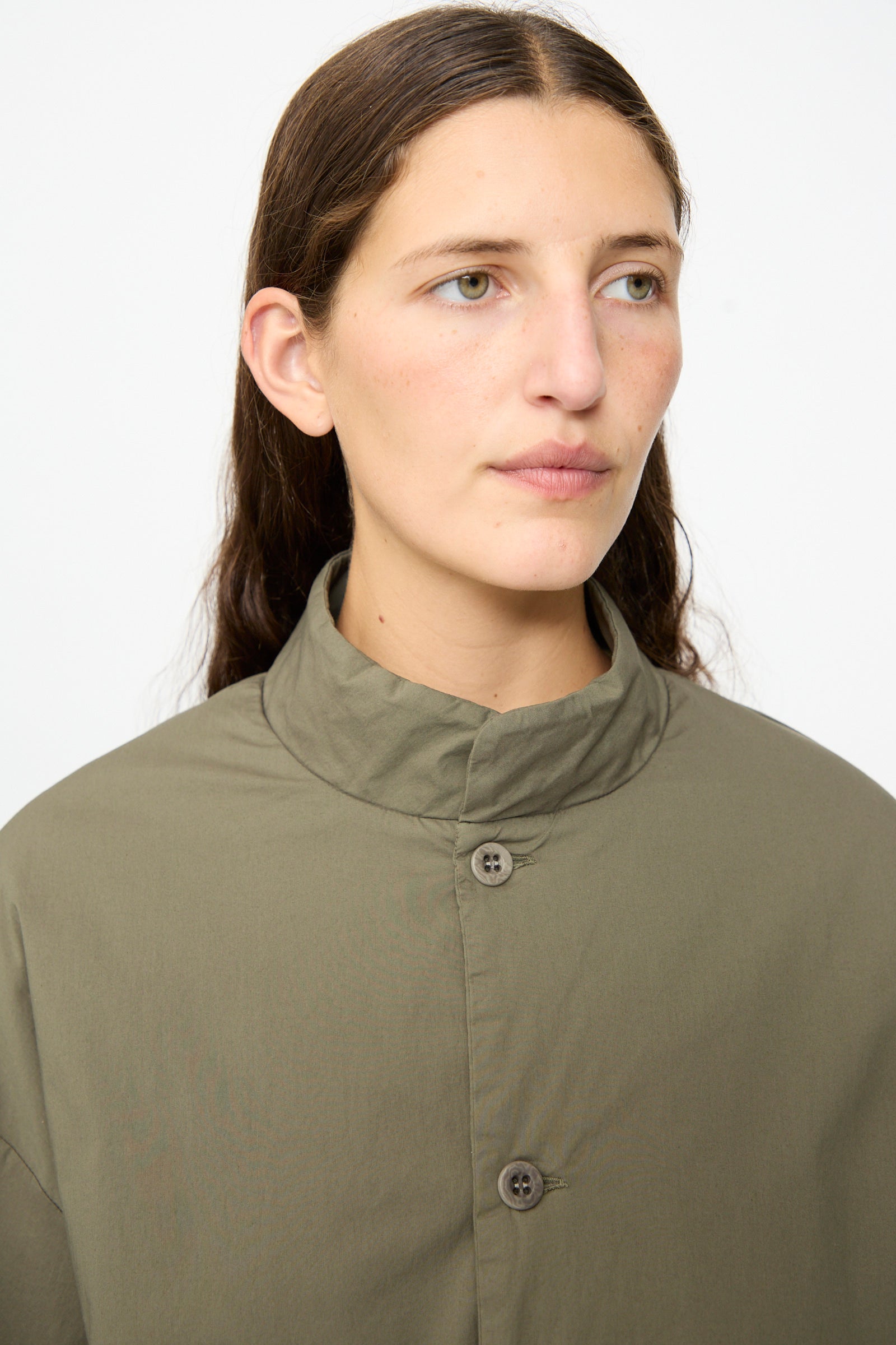 A person with long brown hair, wearing the Labo.Art Ficus Jacket in Lichene—an olive green, button-up collared padded jacket crafted from a cotton blend—looks slightly to the side against a plain white background.