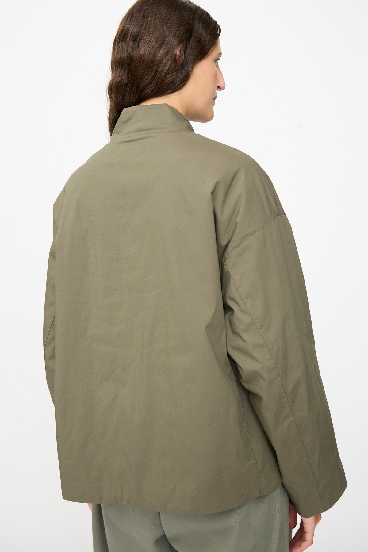 A person with long brown hair, seen from behind against a plain white background, is wearing the Ficus Jacket in Lichene by Labo.Art—an olive green, water-repellent cotton blend jacket—paired with matching pants.