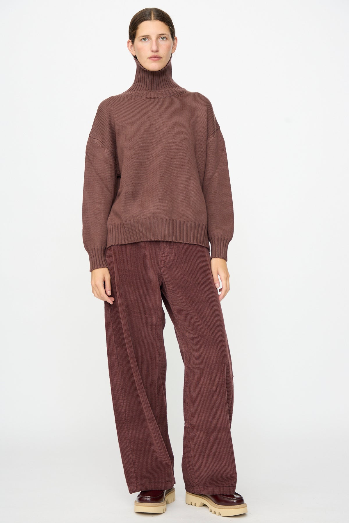 Person standing against a white background wearing the Labo.Art Fiore Sweater in Sasso, paired with wide-leg corduroy pants and beige platform shoes.