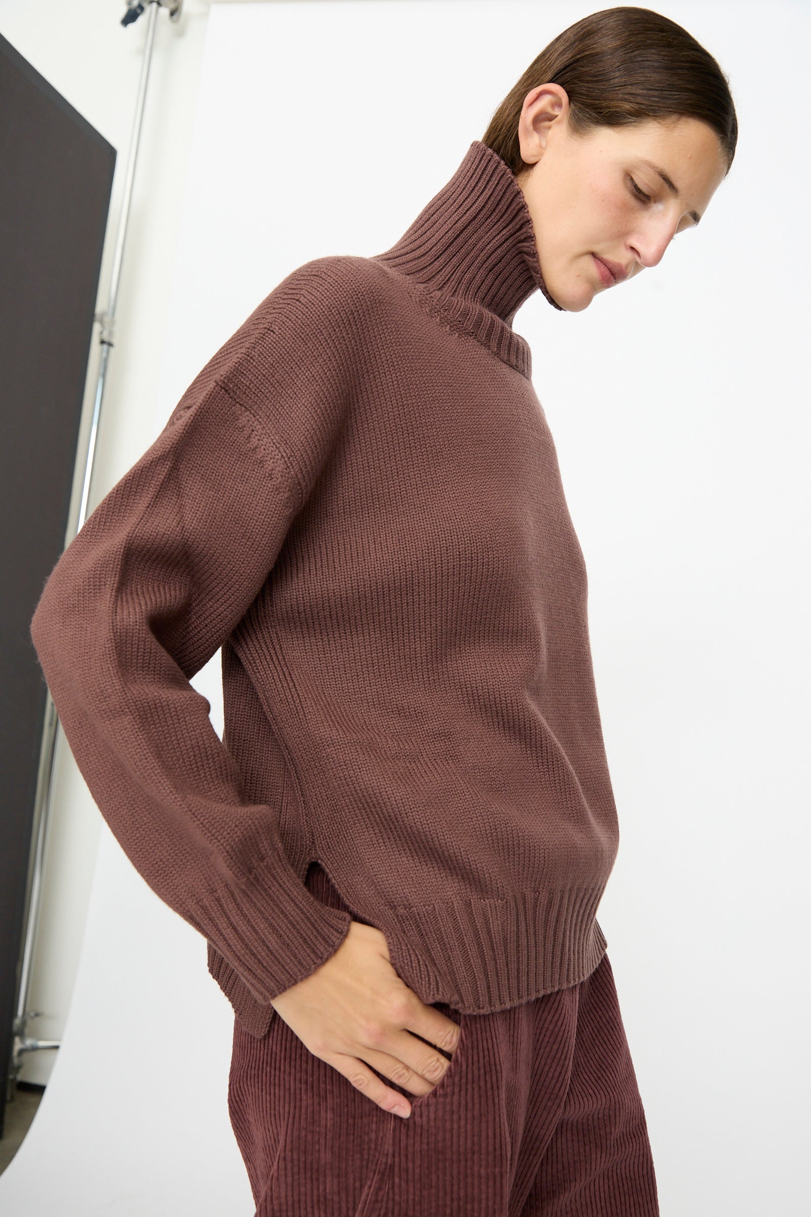 A person stands against a white background, wearing the Labo.Art Fiore Sweater in Sasso—a high-neck brown turtleneck sweater made in Italy—paired with brown corduroy pants, one hand in their pocket and looking downward.