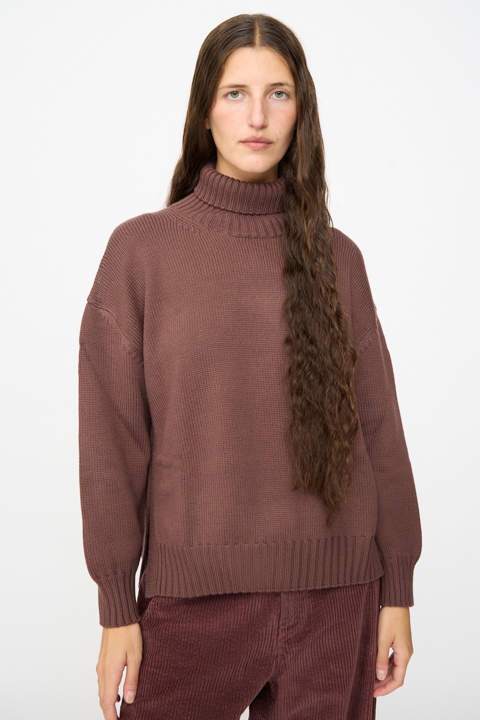 A woman with long brown hair wears the Labo.Art Fiore Sweater in Sasso, a loose brown turtleneck crafted from cashwool knitwear, paired with matching corduroy pants and standing against a plain white background.