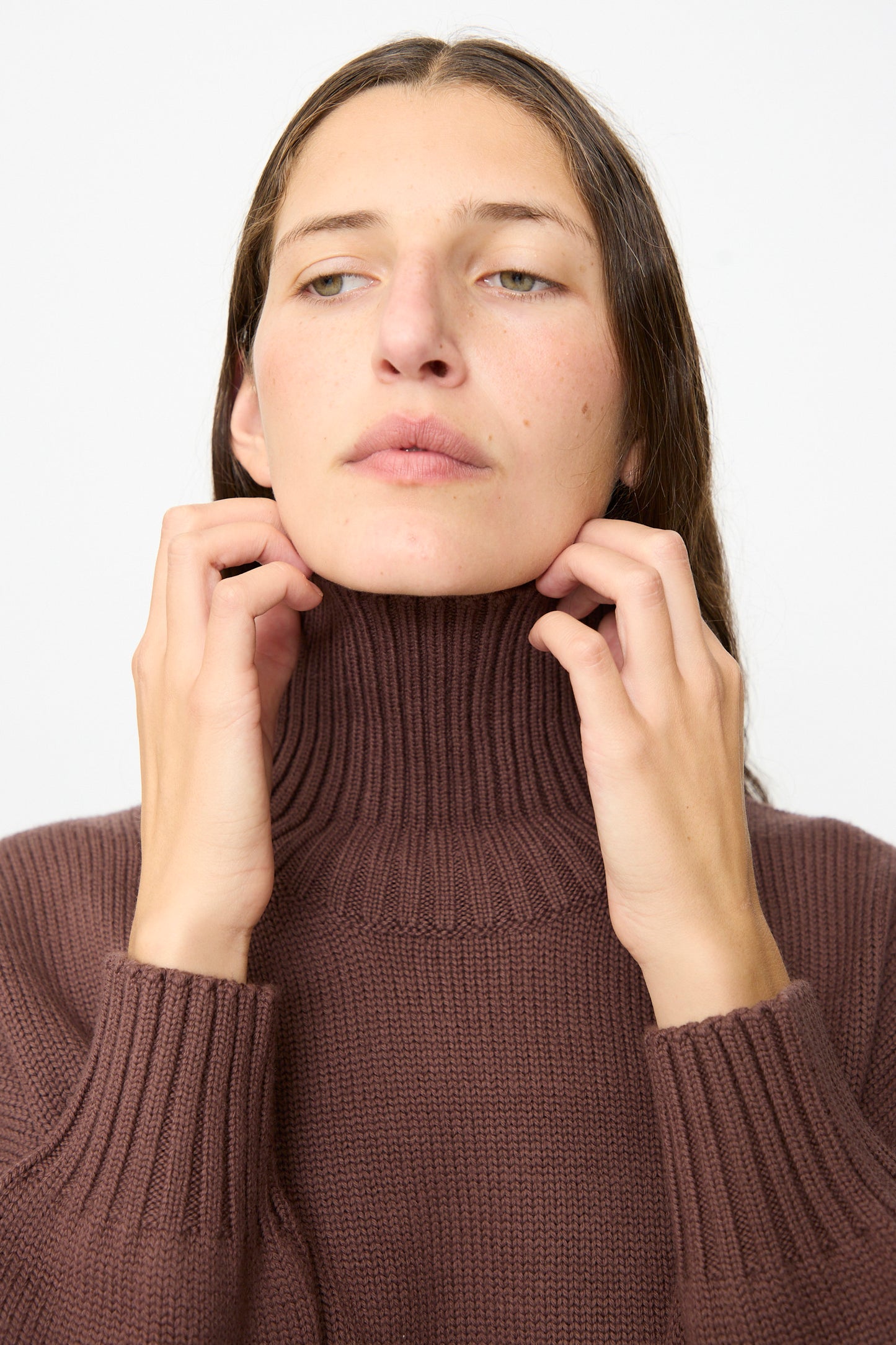 A person is seen wearing the Fiore Sweater in Sasso by Labo.Art—a brown turtleneck made from cashwool knitwear—holding the collar with both hands and gazing slightly upward against a plain background. 